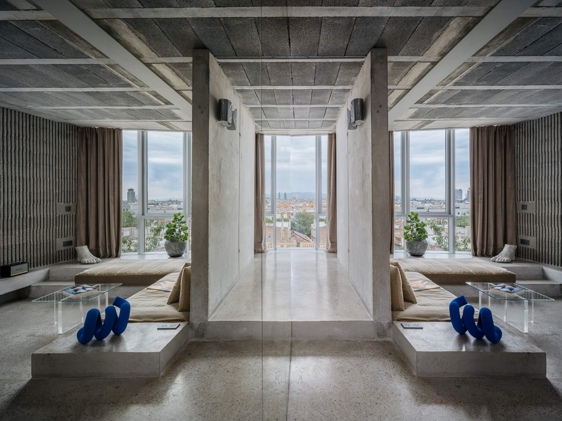 Symmetrical concrete living space with low built-in seating, blue sculptural objects, and large city-facing windows.