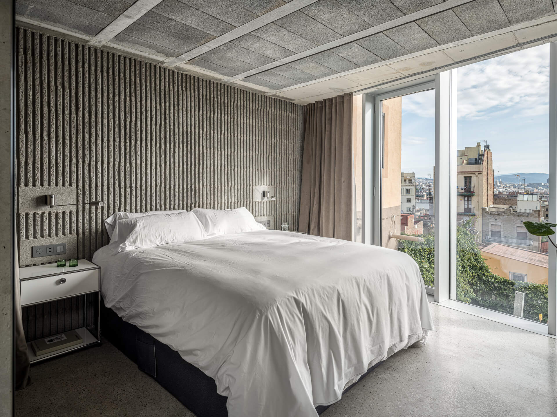 Minimal bedroom with white bedding, textured concrete walls, and a large window overlooking the city.