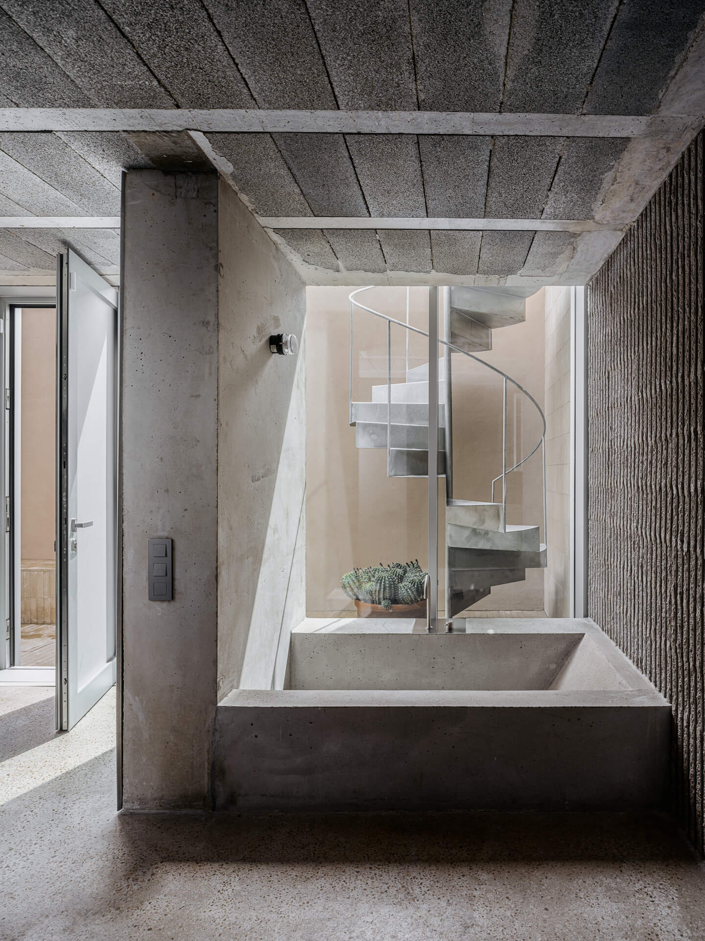 Concrete soaking tub beneath an exposed ceiling with a white spiral staircase in the background.