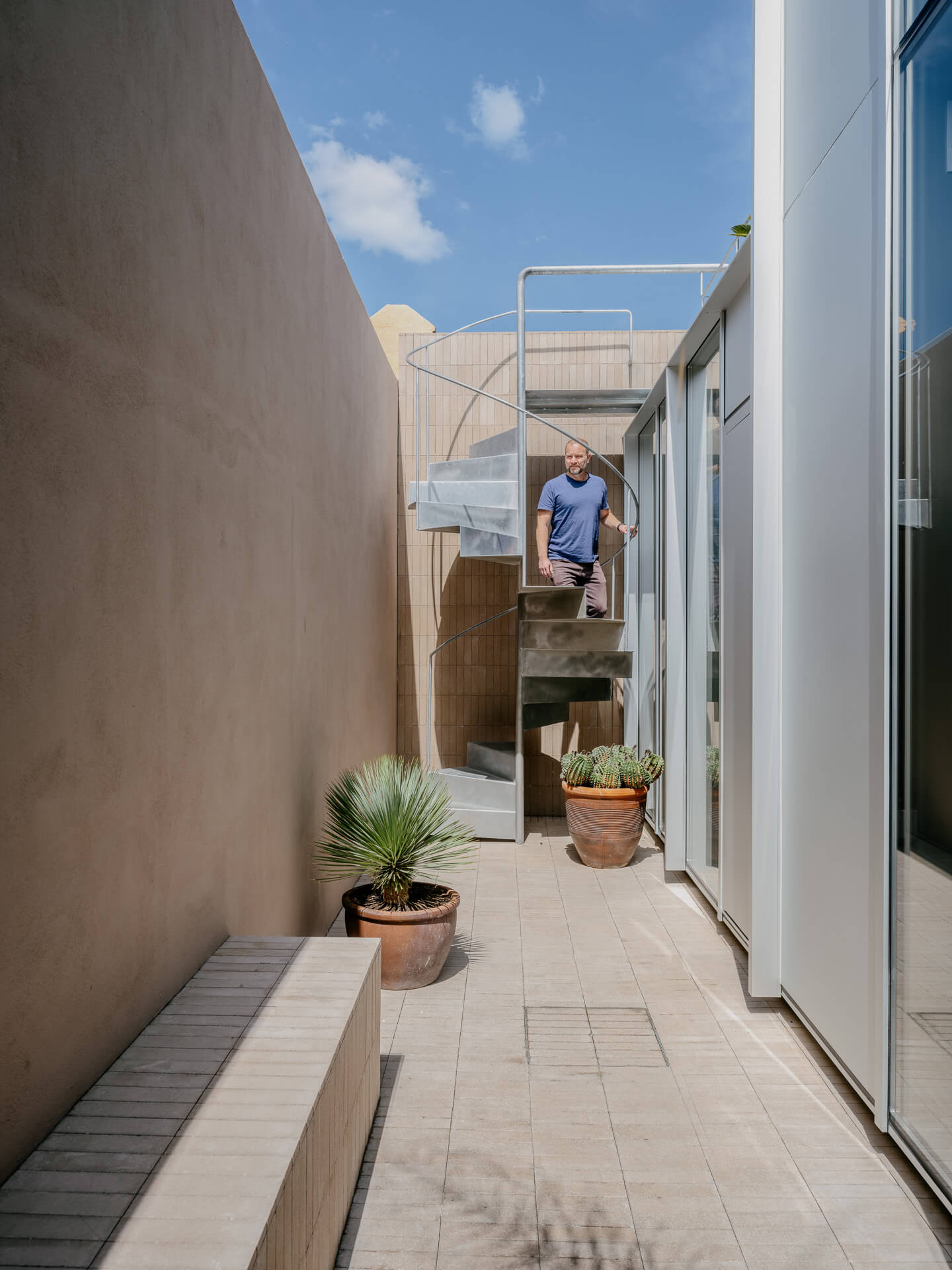 Narrow outdoor terrace lined with potted plants, with a person standing on steps at the far end.