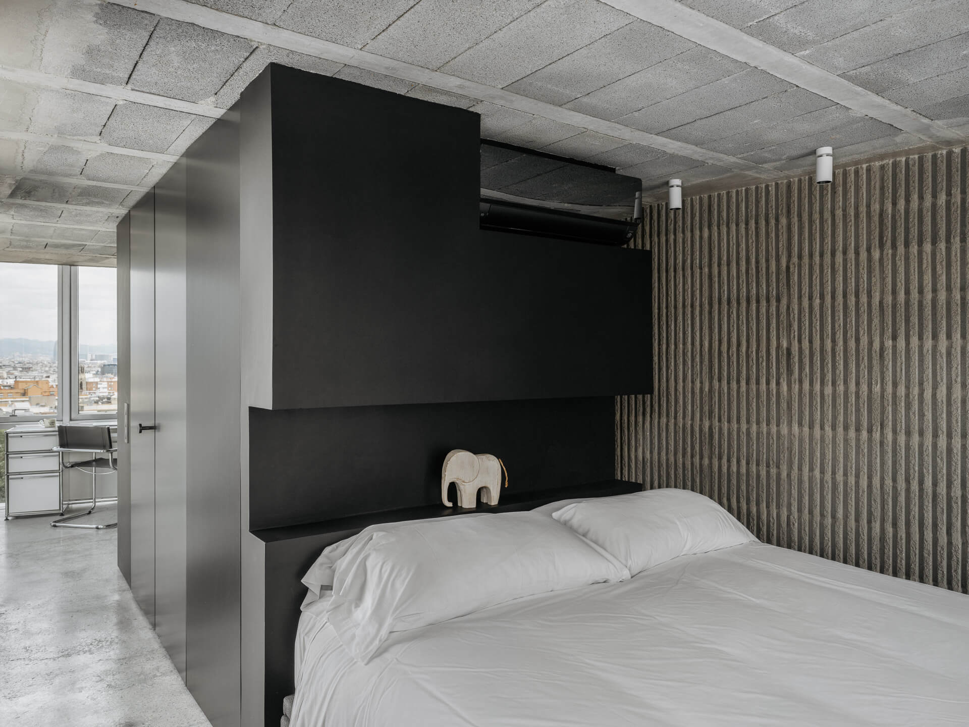 Bedroom with white bedding beneath a dark architectural volume and textured concrete walls.
