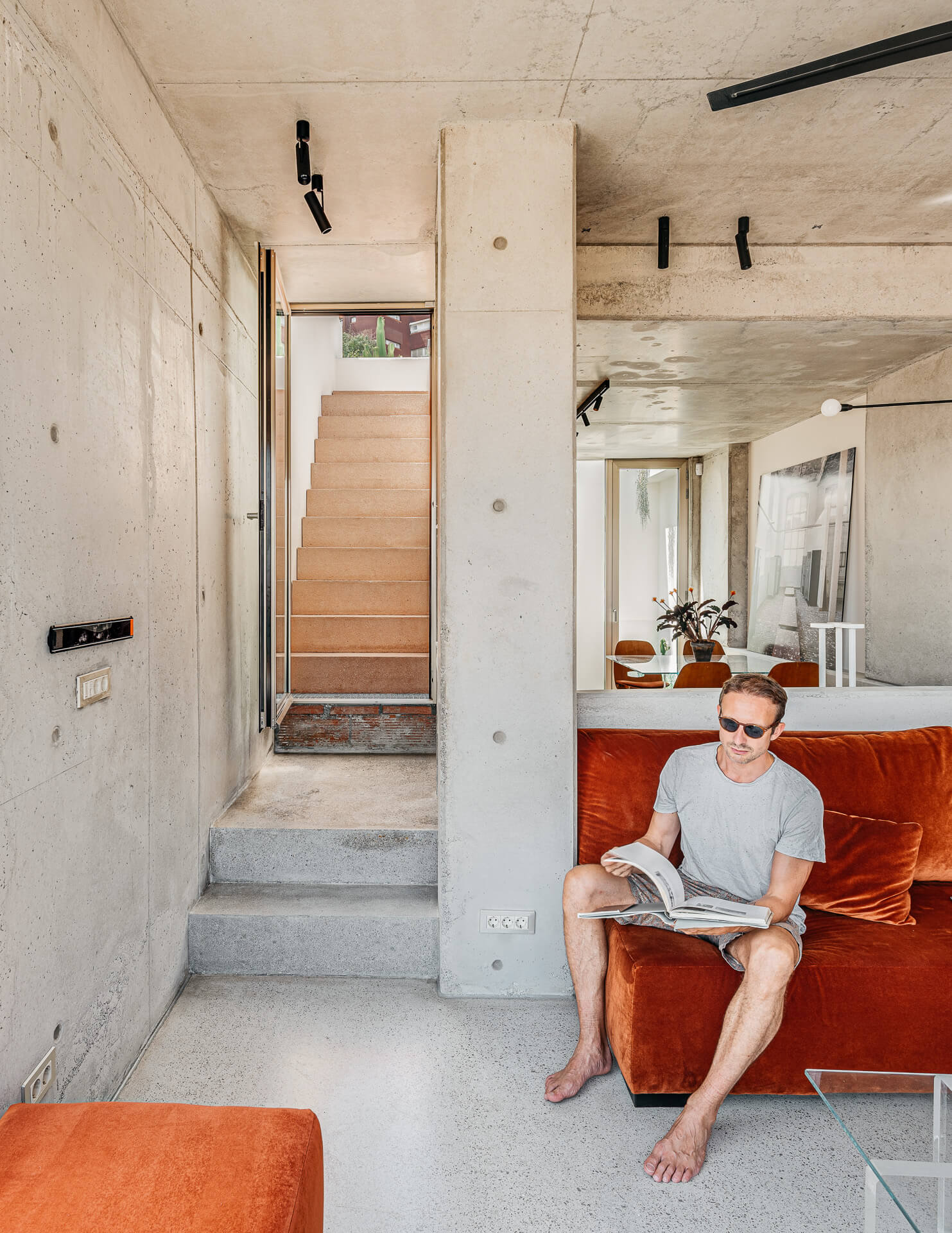 Homeowner Christian Schallert seated on a rust-colored sofa in an open concrete interior with a staircase beyond.