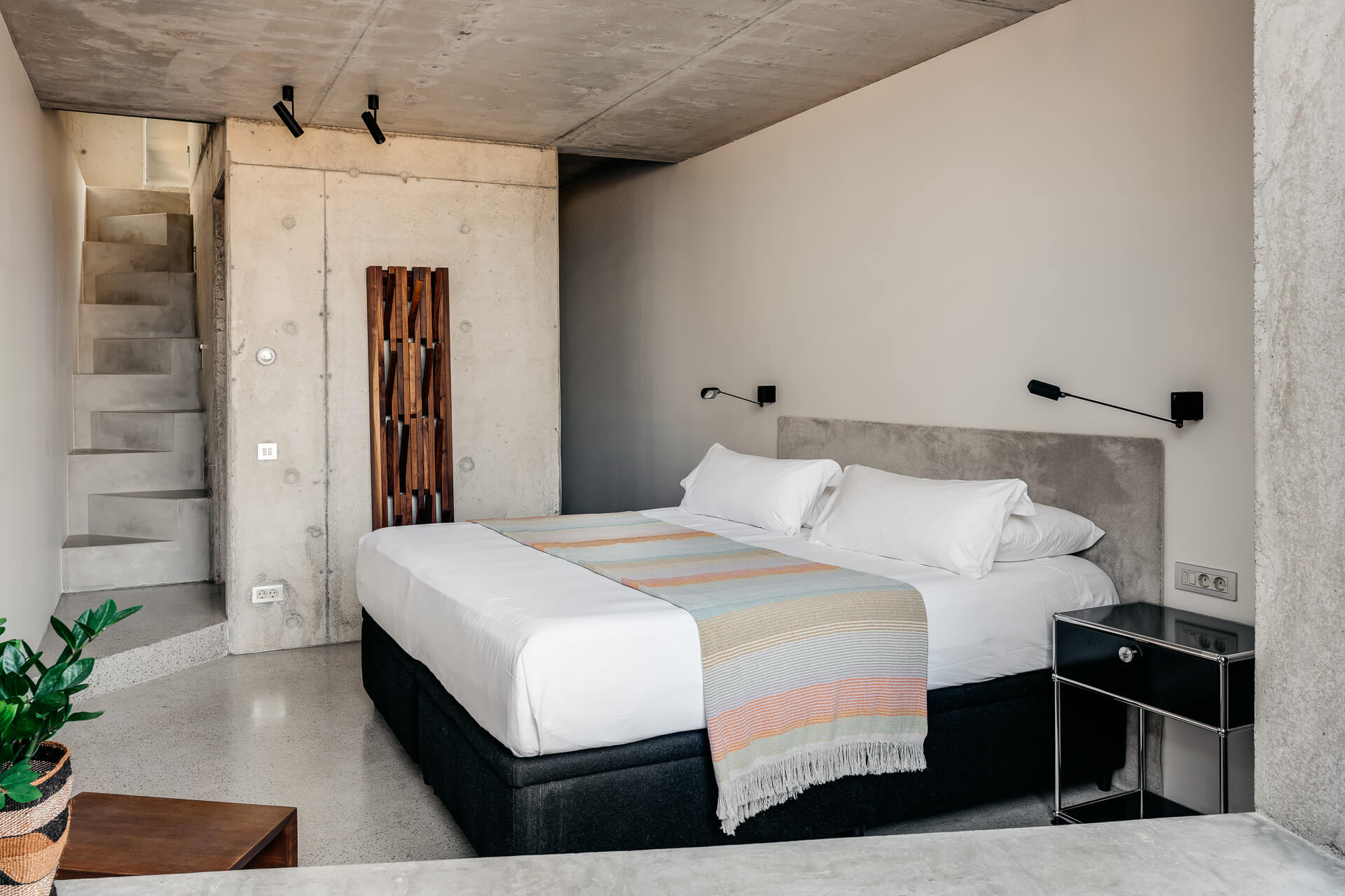 Minimal bedroom with raw concrete walls, white bedding, open shelving, and a striped throw