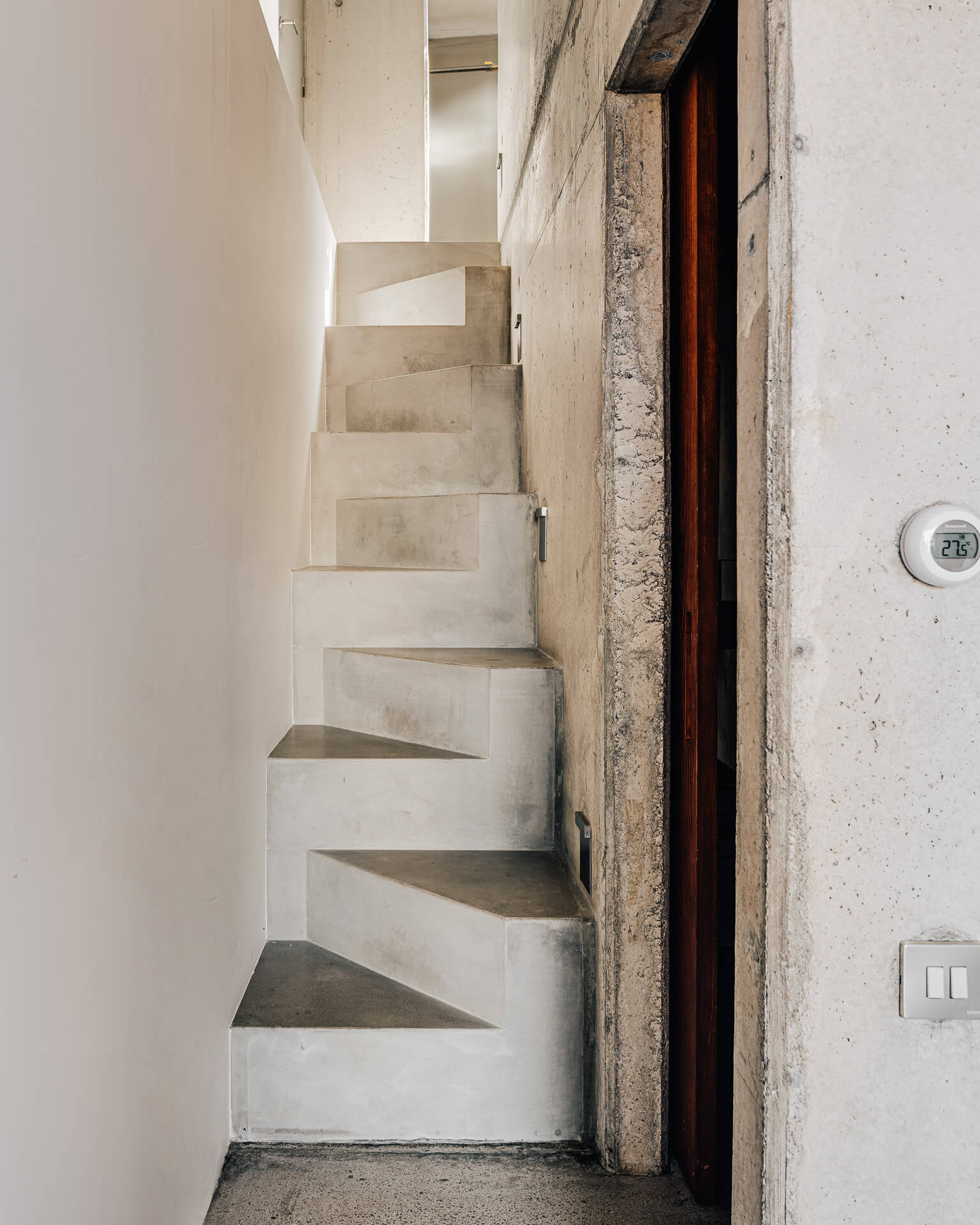 Sculptural concrete staircase rising beside a narrow doorway and textured concrete wall