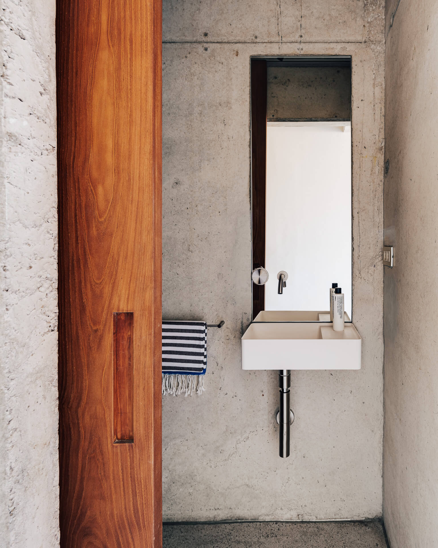 Compact white sink and mirror set against a raw concrete wall beside a wood door.