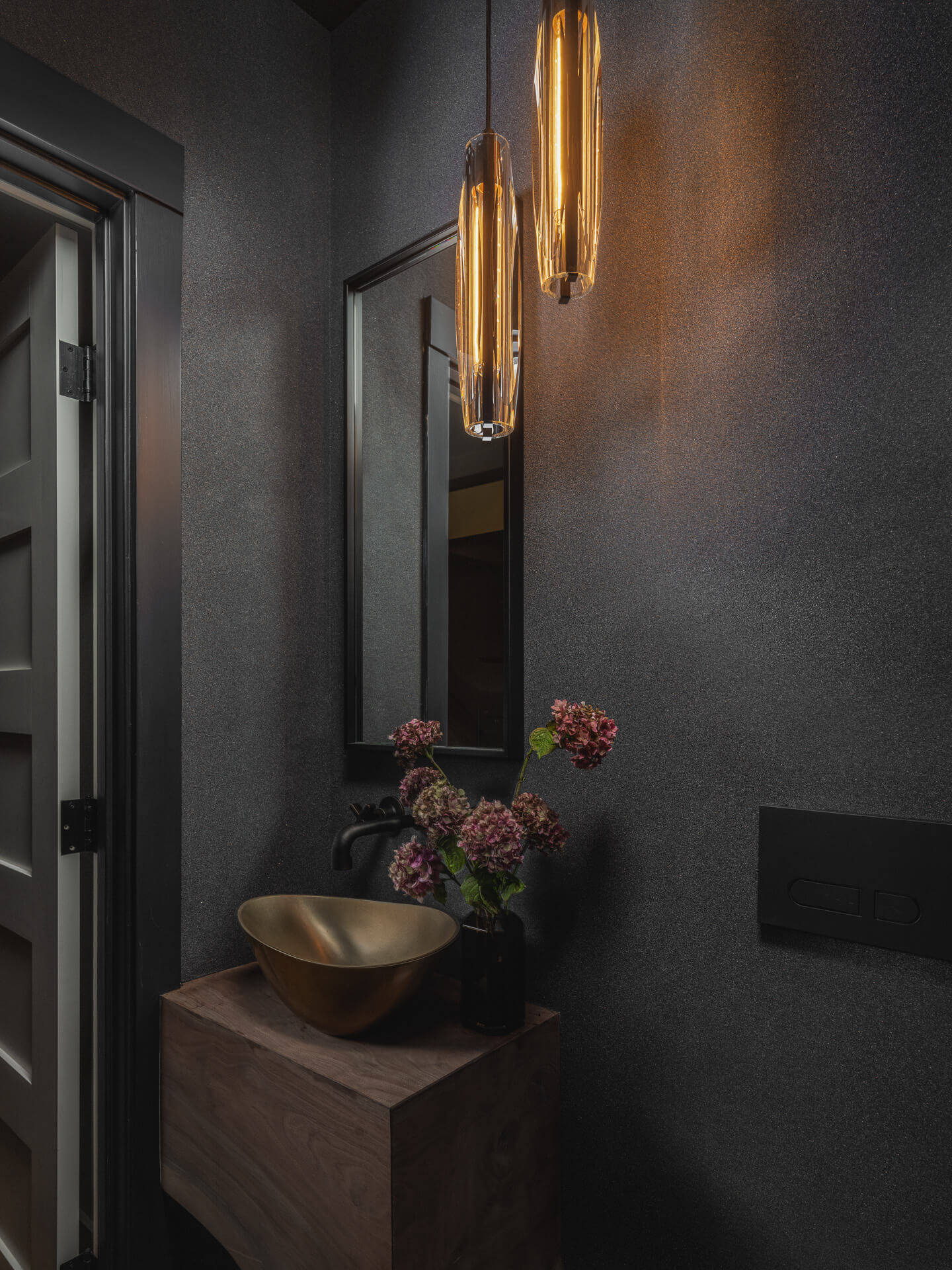 Dark moody powder room with textured walls, glowing brass sconces and a small wood console with flowers.