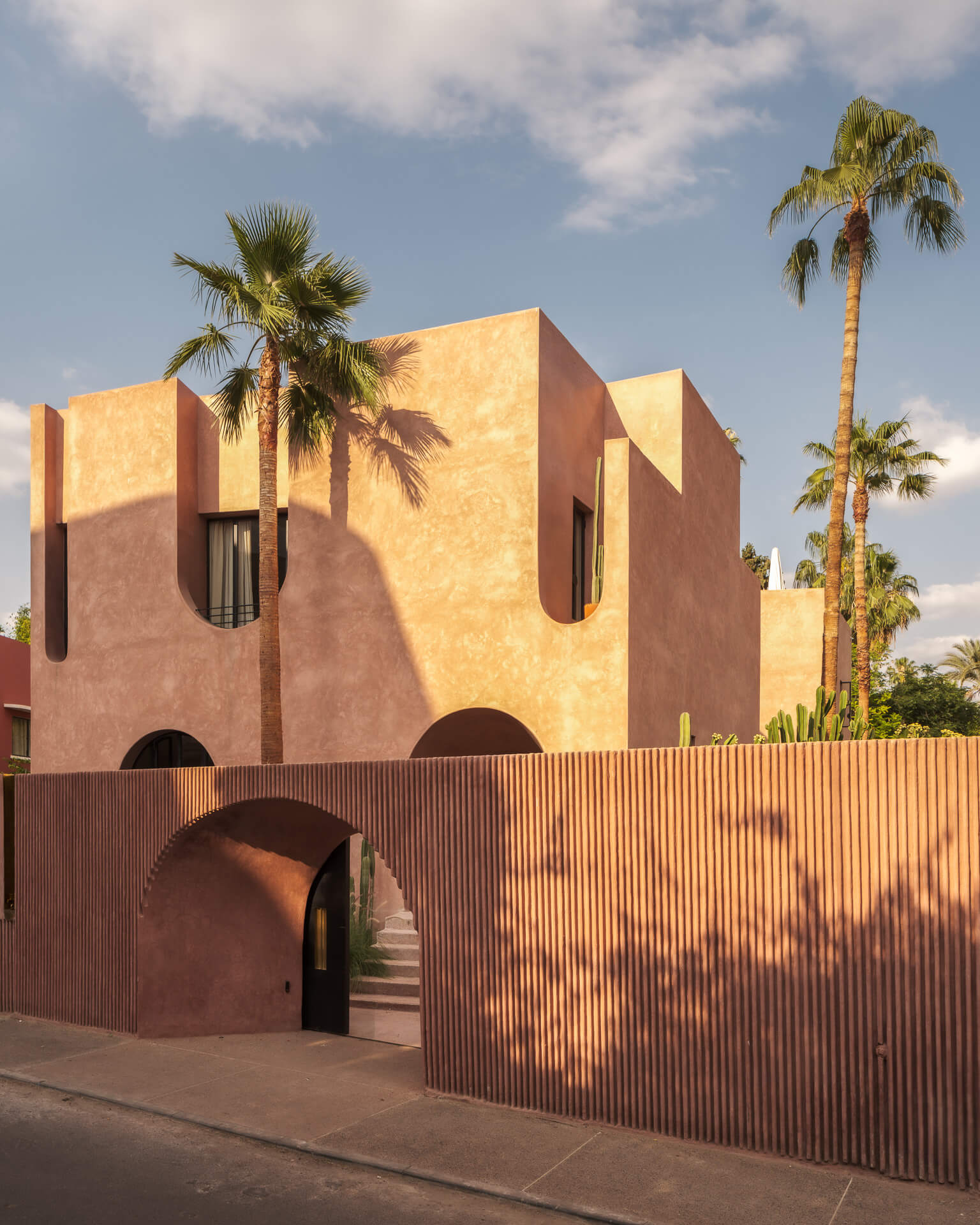 Pink stucco hotel with sculptural arches and tall palm trees beneath a blue sky.