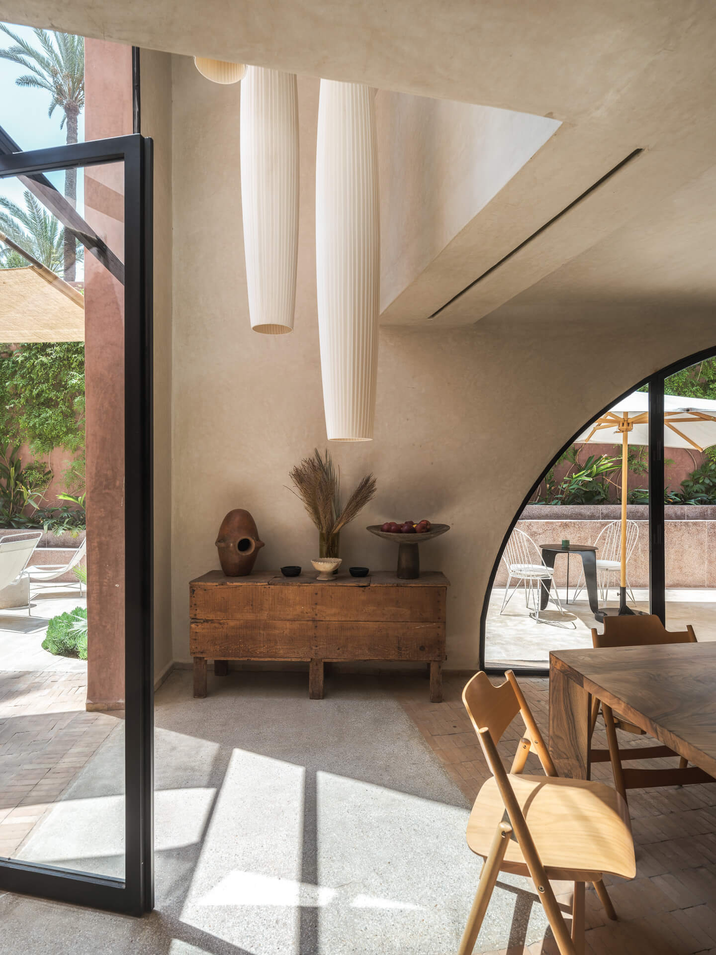 Sunlit dining area with wood table and chairs, curved plaster walls, and glass doors opening outdoors.