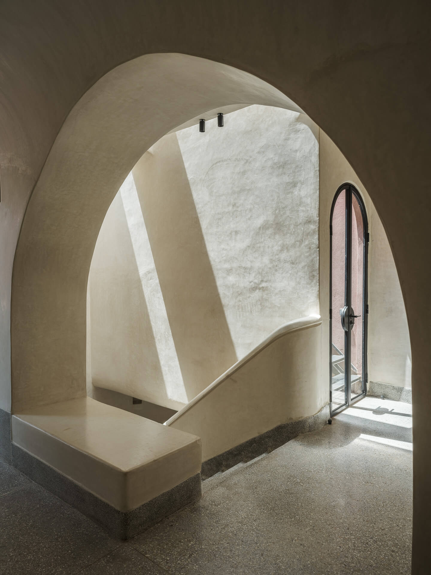 Curved plaster staircase and landing illuminated by sunlight from a tall arched window.