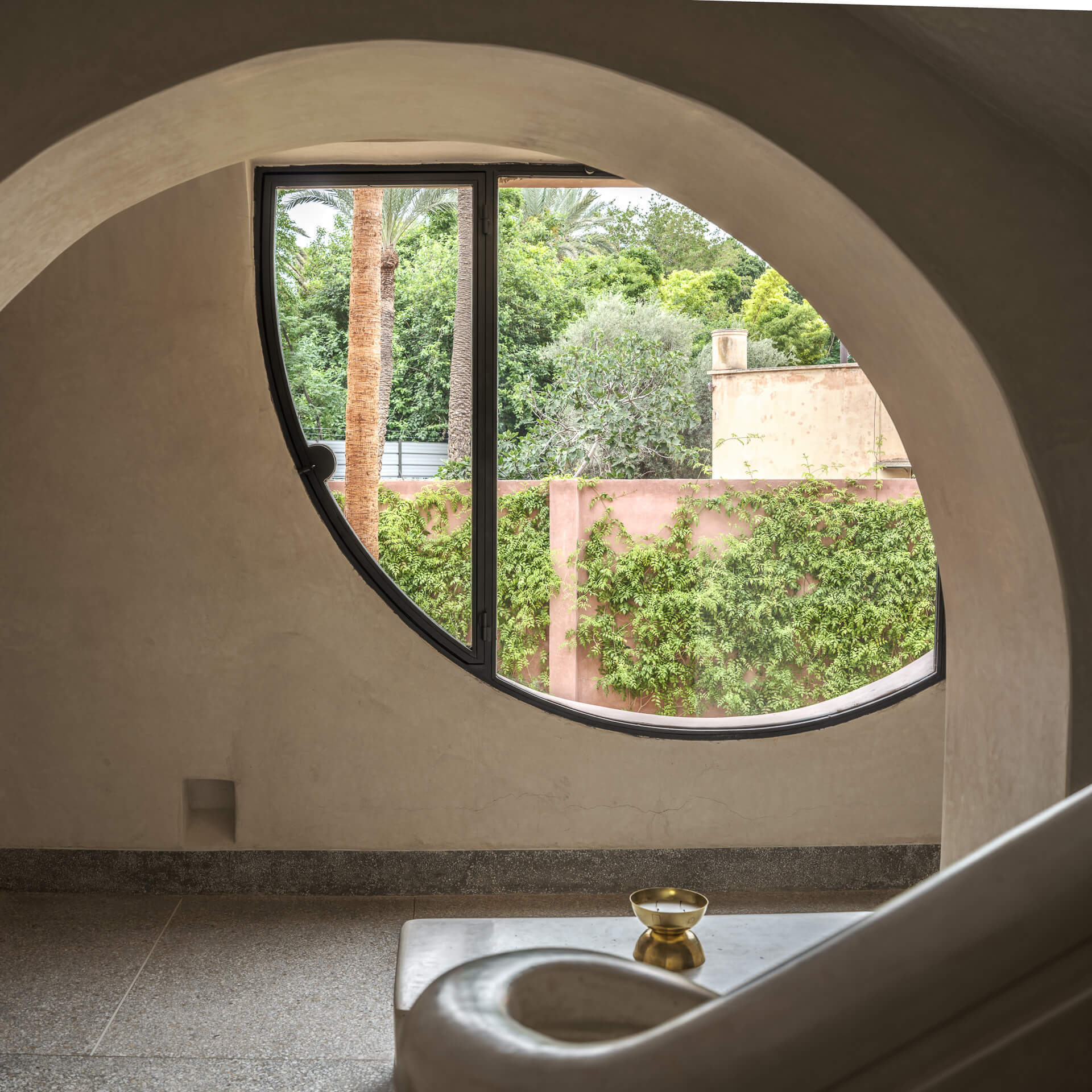 Organic rounded window in a pale plaster wall framing palms and garden greenery.