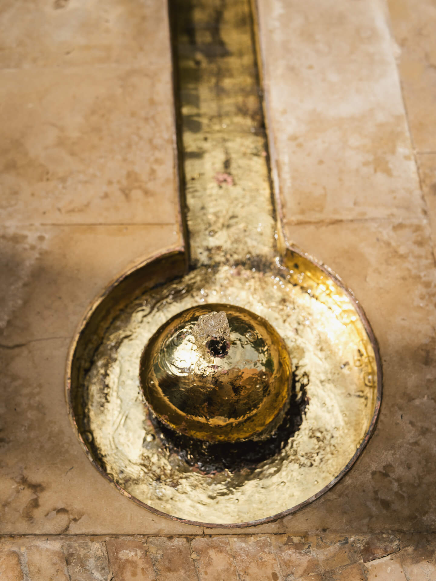 Close-up of a circular aged-brass fountain set into a pale stone surface.