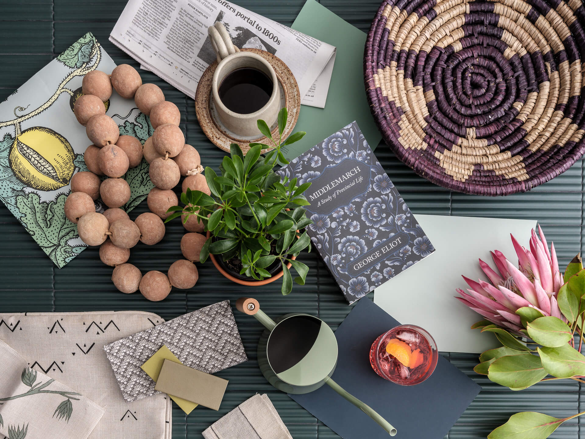 Overhead flat lay of woven baskets, ceramic vessels, fabric samples, flowers and decorative objects on a muted green surface.