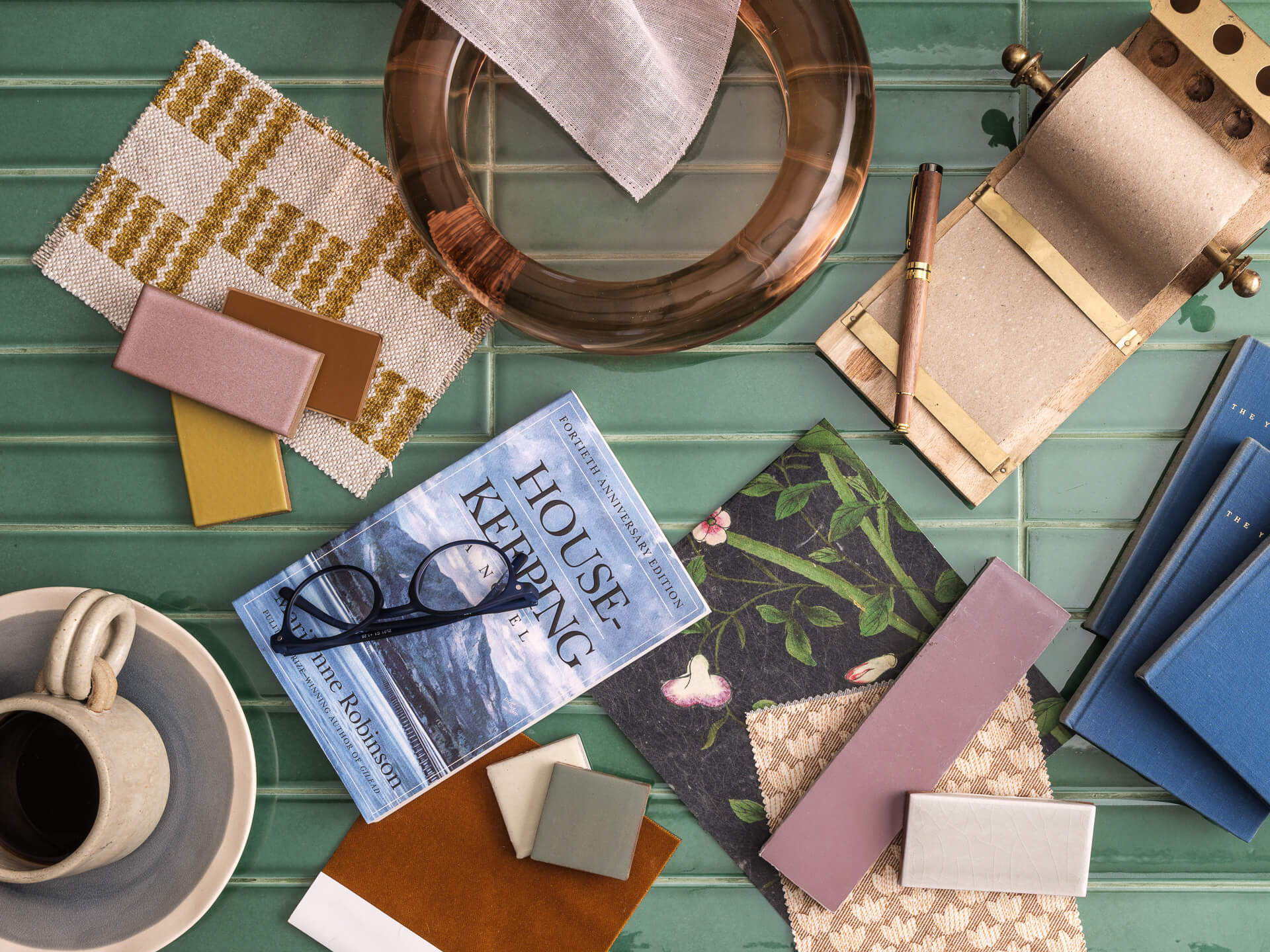 Layered flat lay of design books, textile samples, trays, ceramics and geometric objects in warm muted colors.