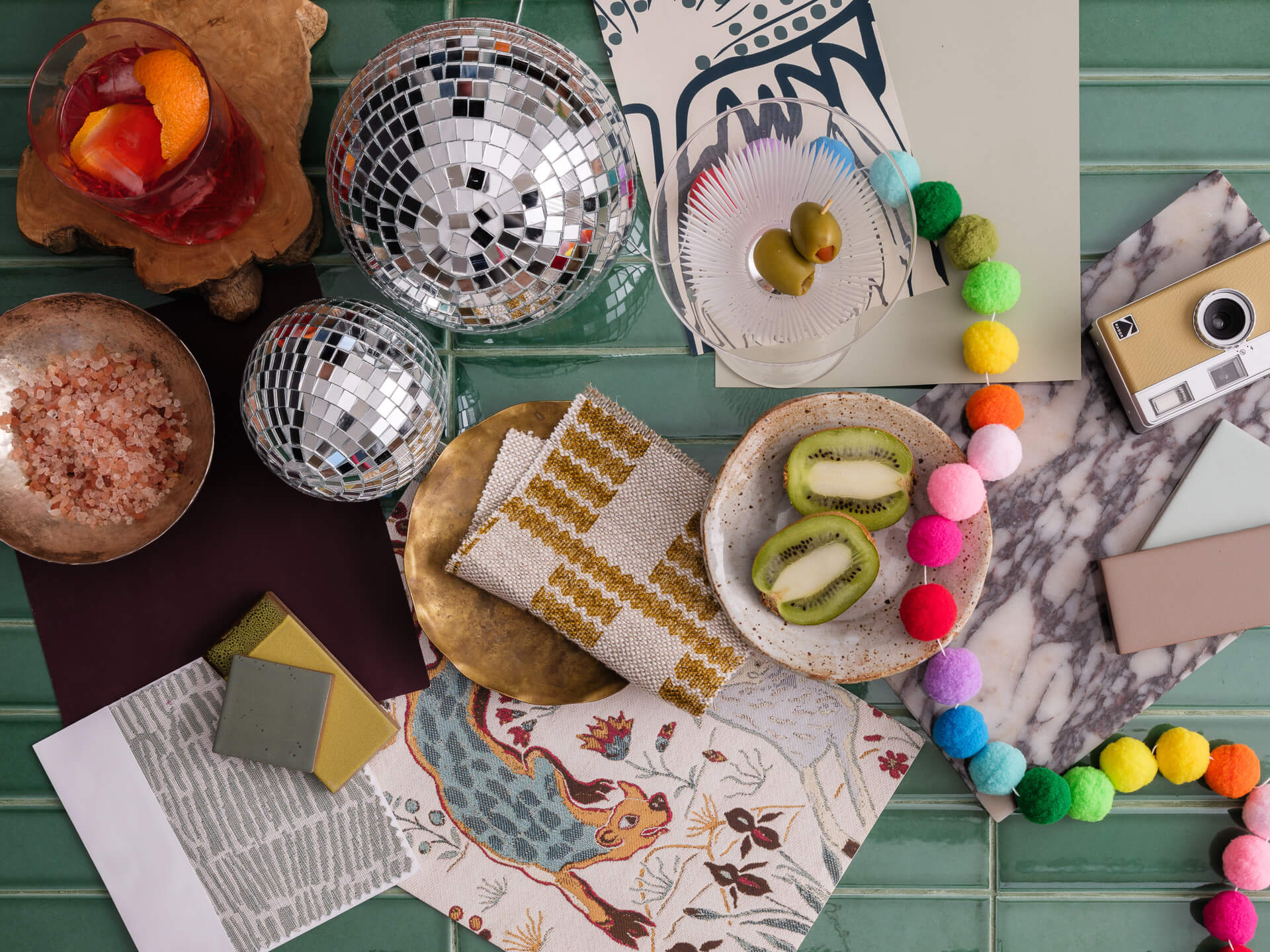 Colorful tabletop arrangement with fruit, patterned dishes, glassware, textiles and a bright beaded necklace.