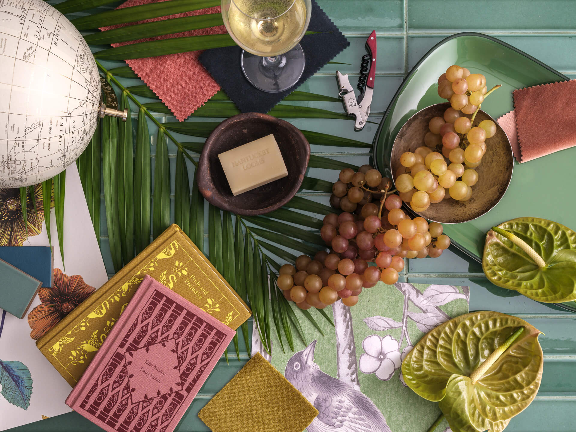 Overhead composition of grapes, sculptural fruit, books, textiles, ceramics and decorative objects on a green table