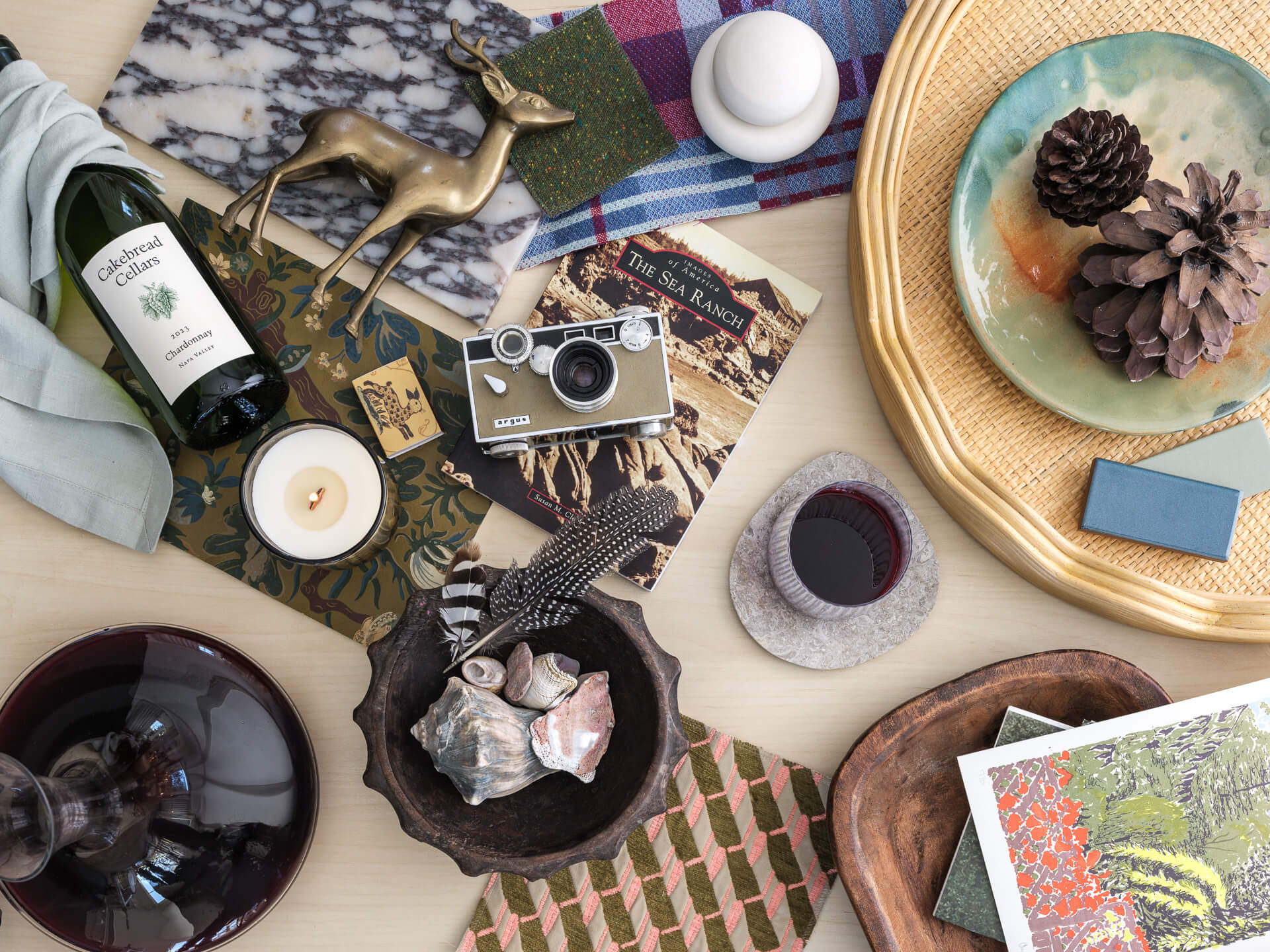 Layered flat lay with wine bottle, pinecones, vintage camera, textiles, books and decorative objects
