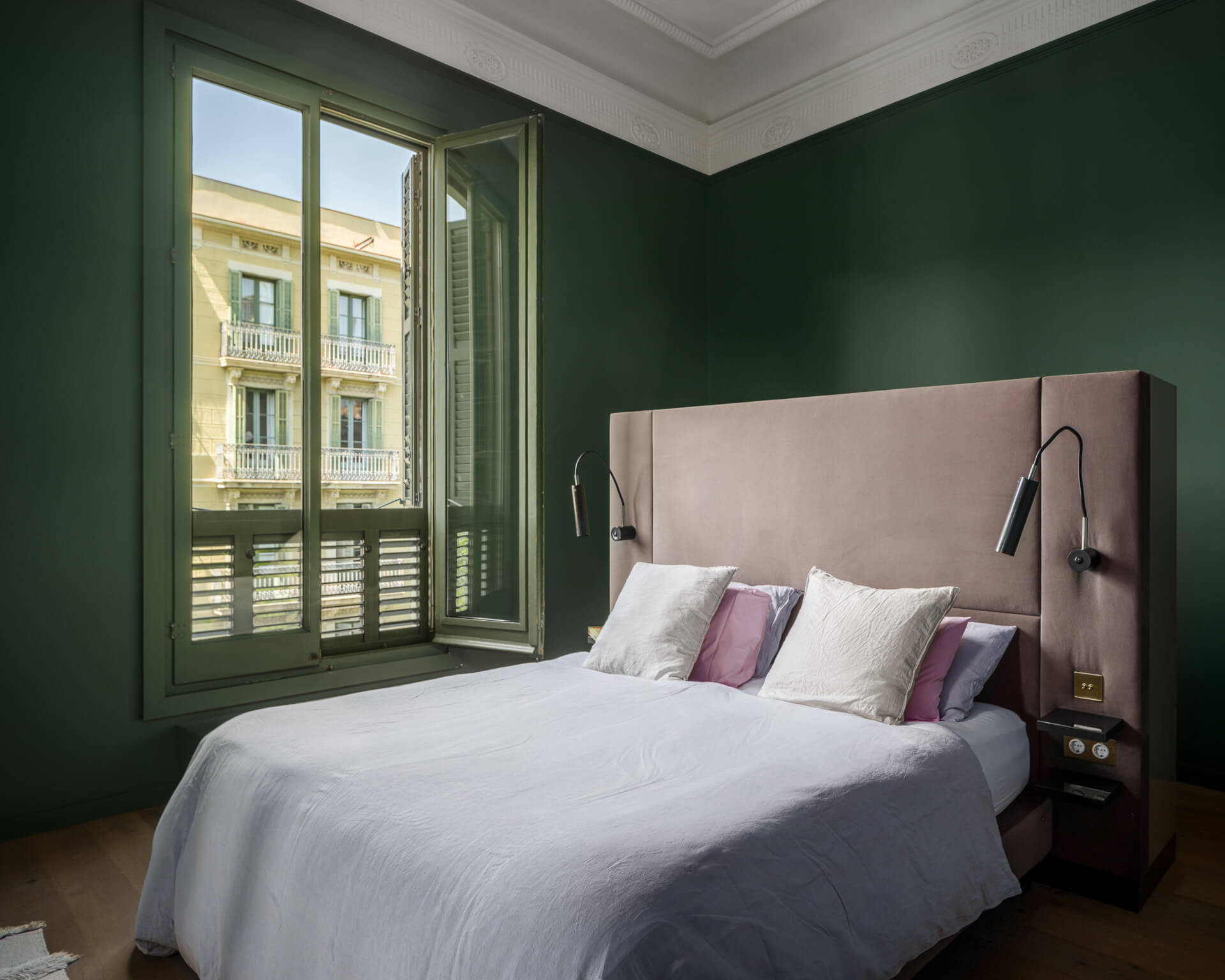 Serene bedroom with deep green walls, blush upholstered headboard, white bedding, and tall shuttered windows.