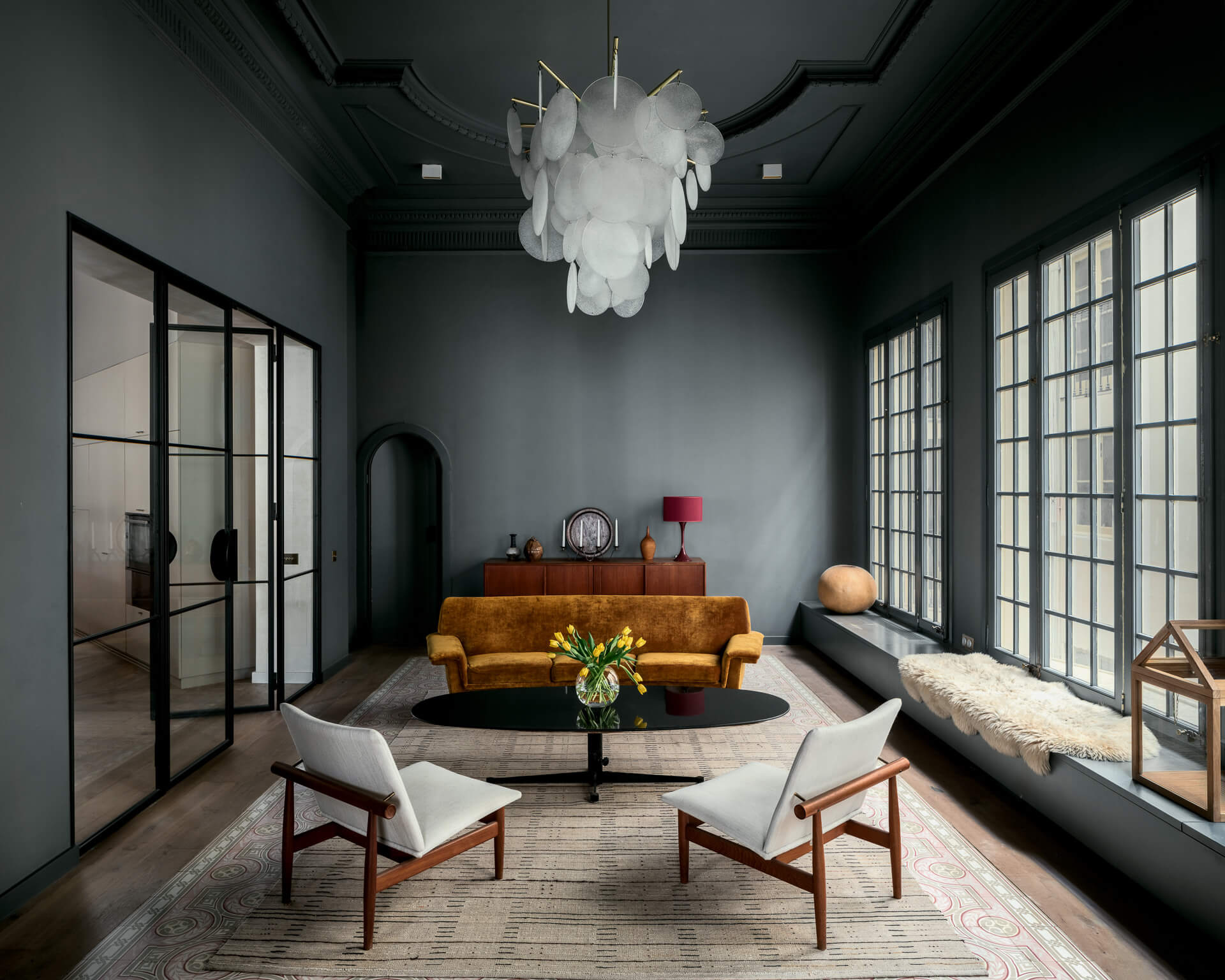 Dark green sitting room with caramel sofa, white lounge chairs, window bench, and sculptural chandelier.