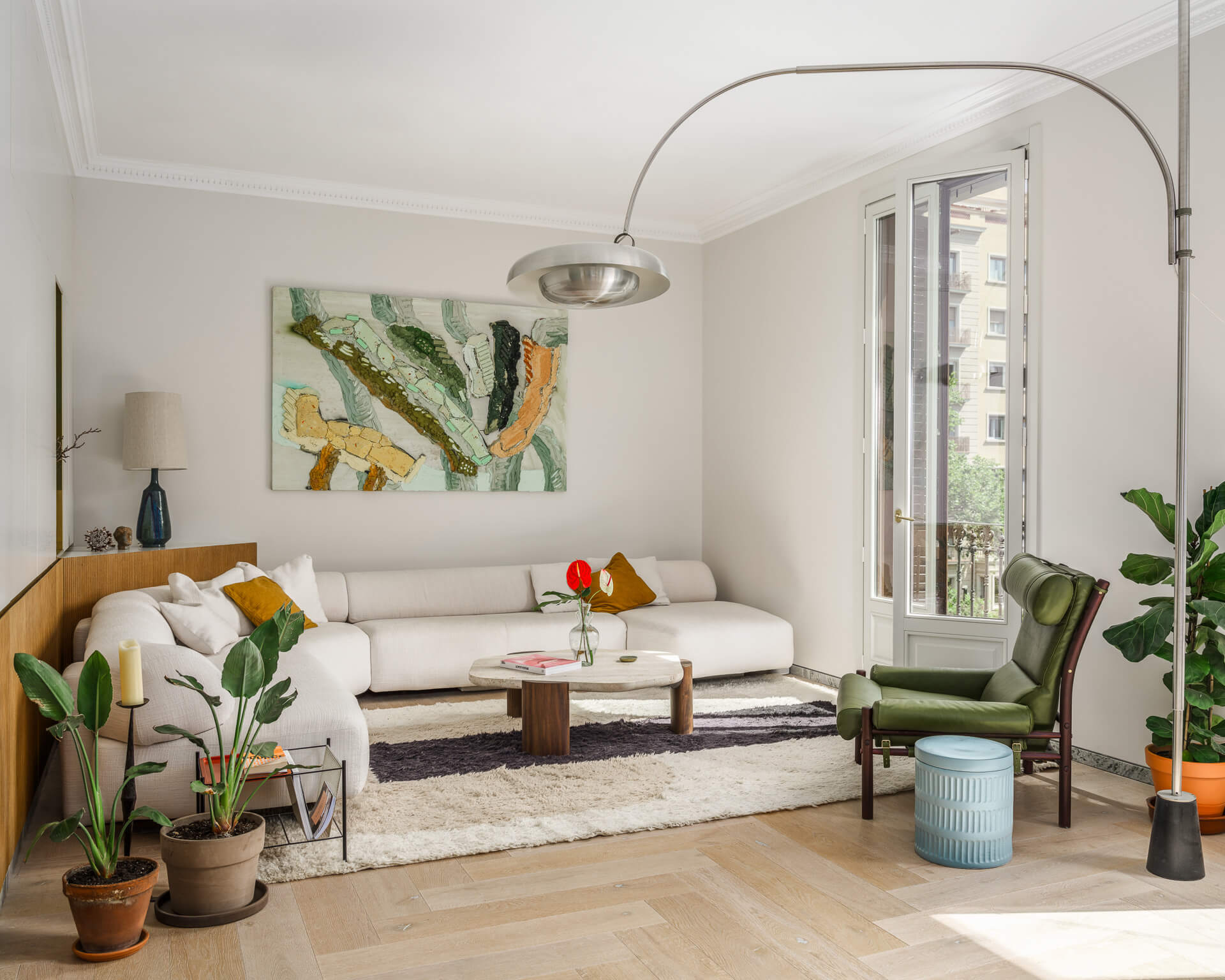 Bright living room with white sectional sofa, green lounge chair, potted plants, colorful artwork, and tall balcony doors.