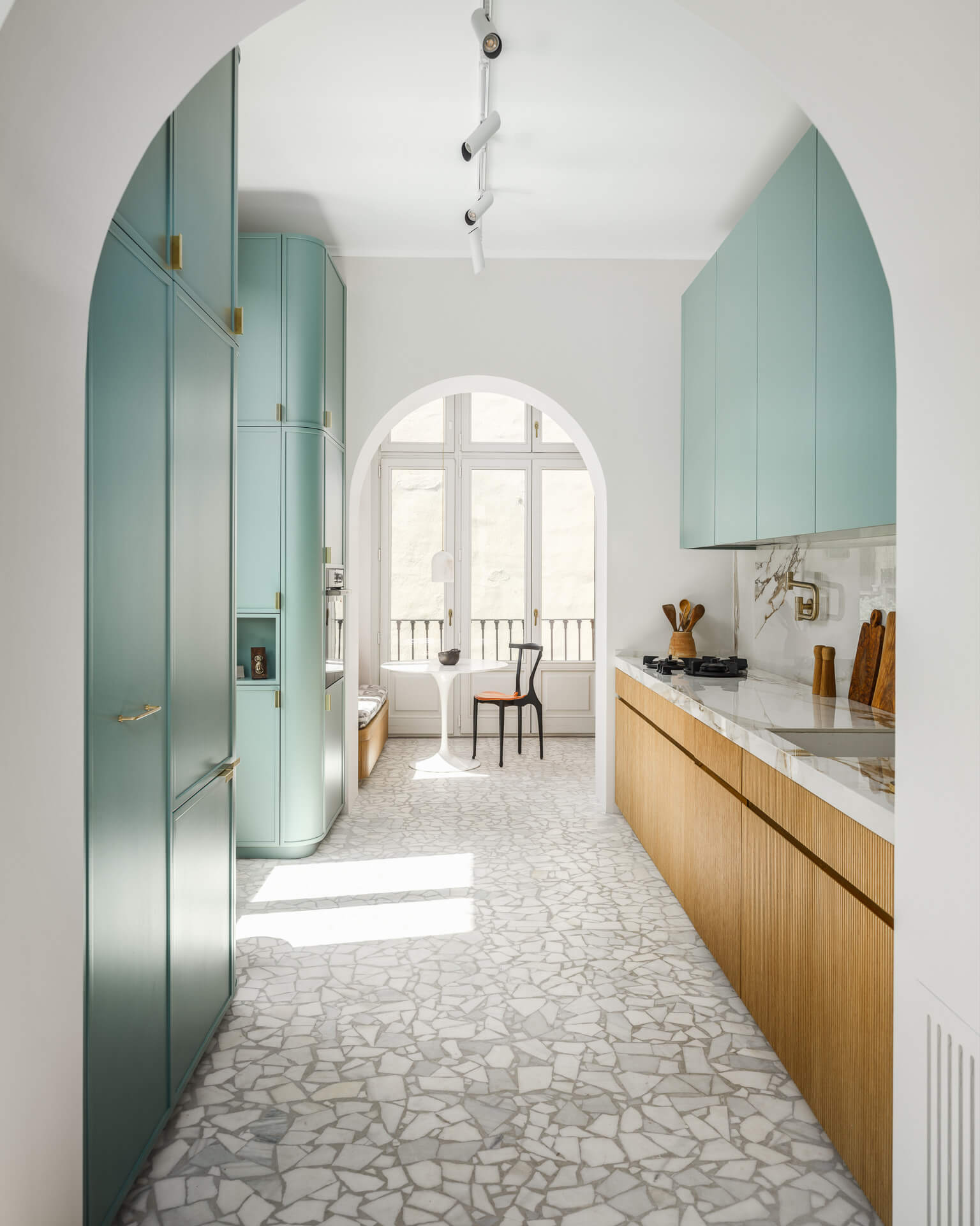 Long kitchen with pale green cabinetry, wood lower cabinets, stone flooring, and an arched window at the far end.