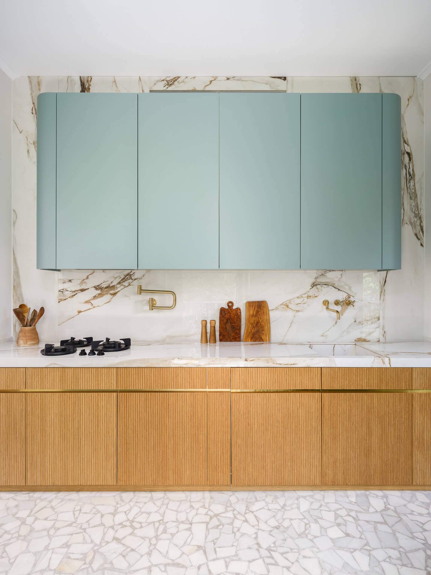 Kitchen detail with pale green upper cabinets, ribbed wood lower cabinetry, and dramatically veined stone backsplash.