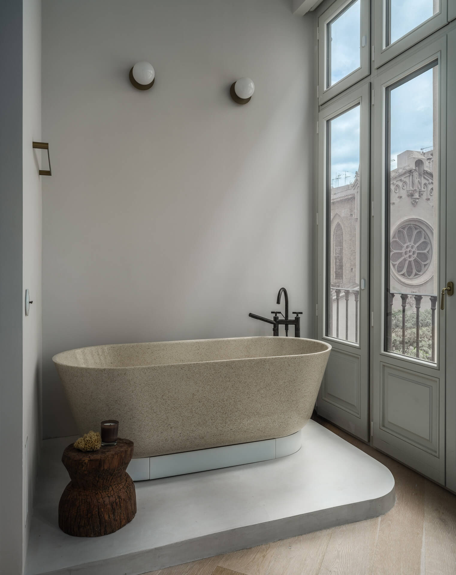 Freestanding stone bathtub beside tall windows overlooking neighboring architecture.