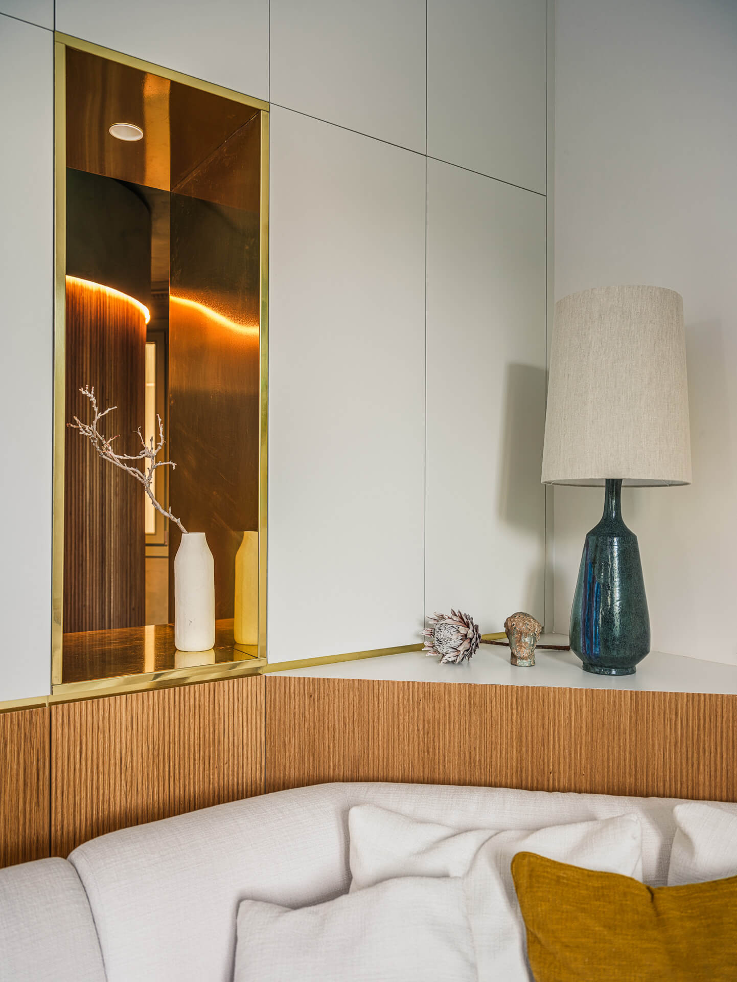 Built-in white cabinetry above a curved cream sofa, with warm wood paneling, display niche, and blue table lamp.