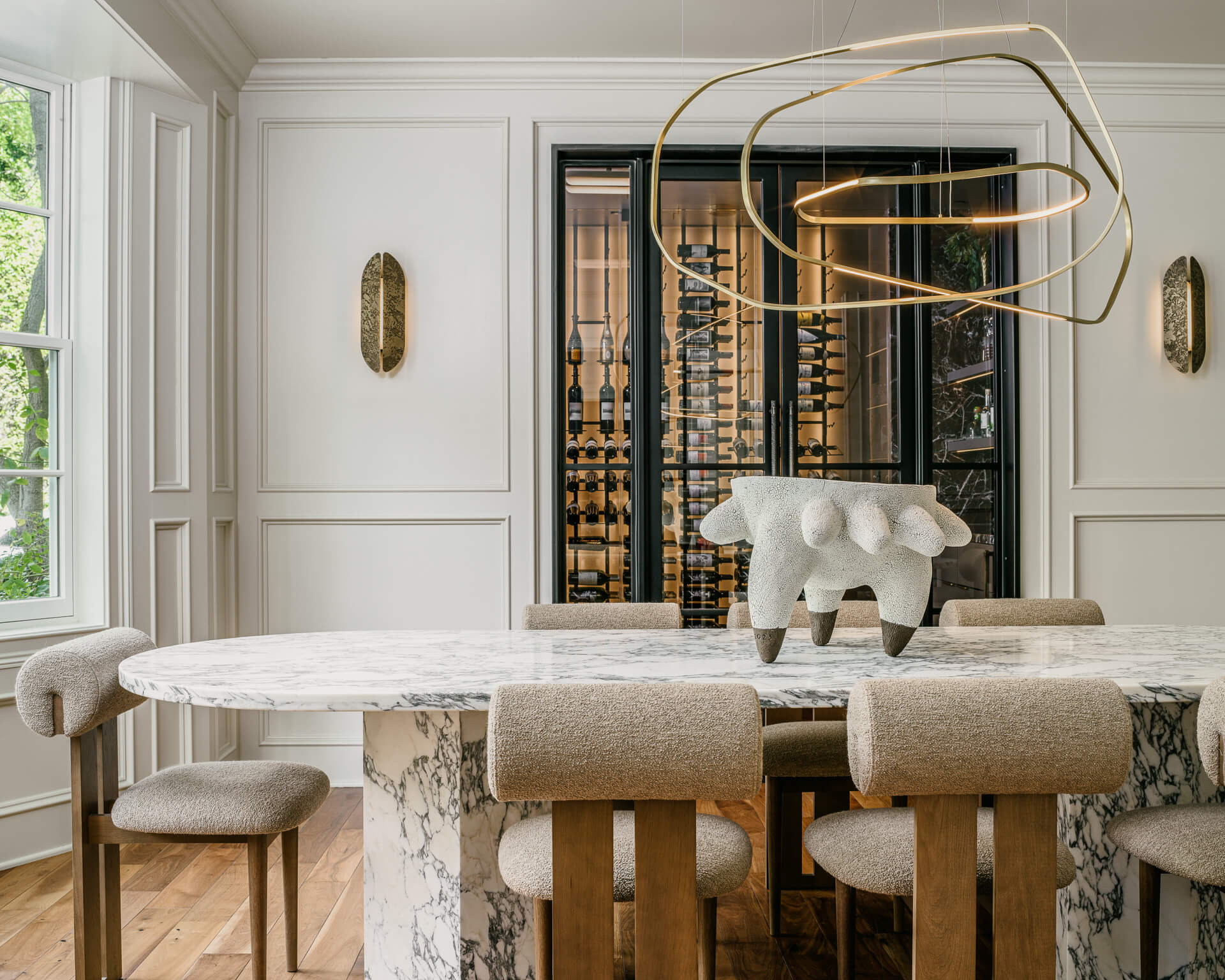 Elegant dining room with marble table, upholstered chairs, sculptural brass chandelier, and glass-front wine storage.