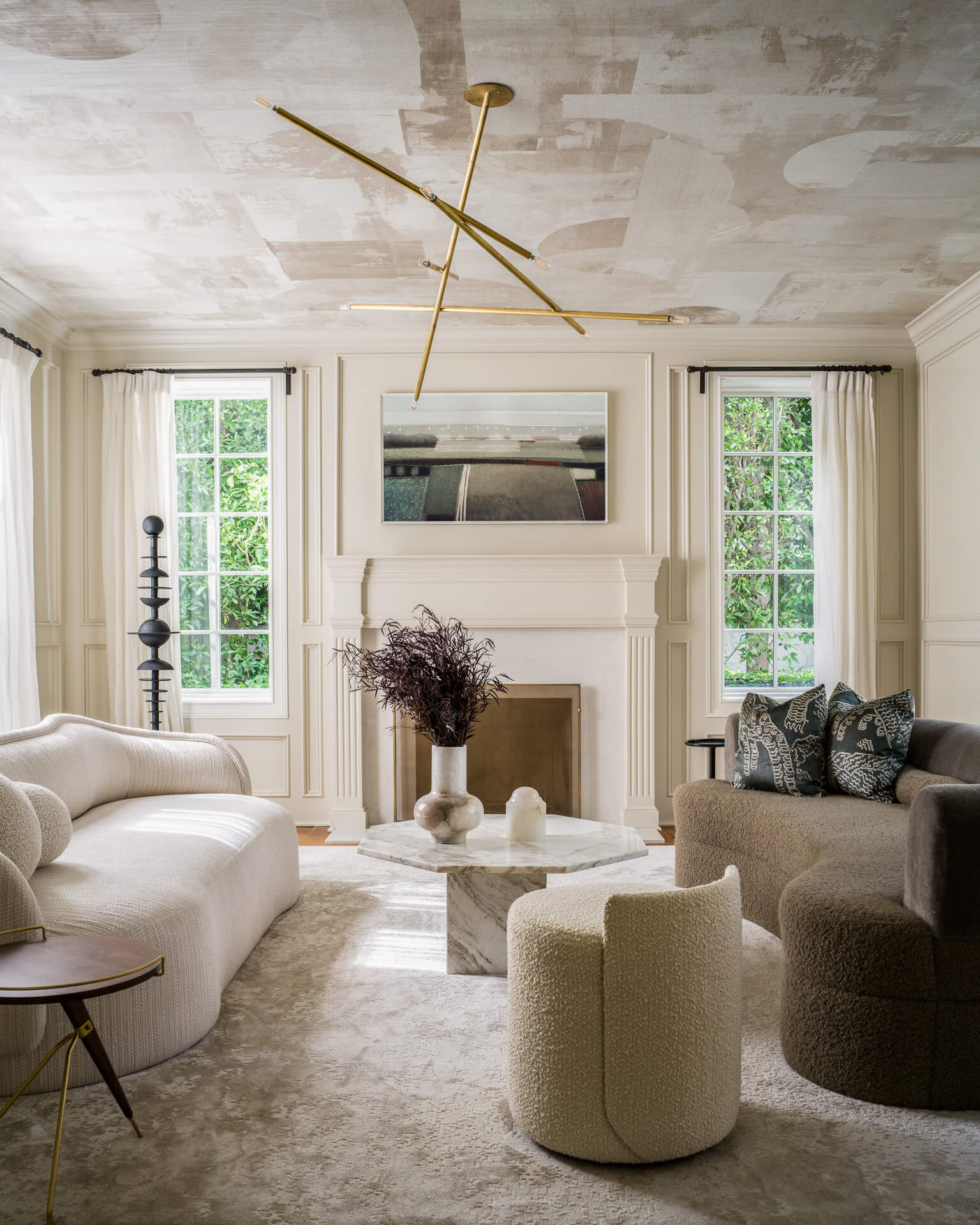 Neutral living room with curved seating, fireplace, textured ceiling, and tall windows overlooking greenery.