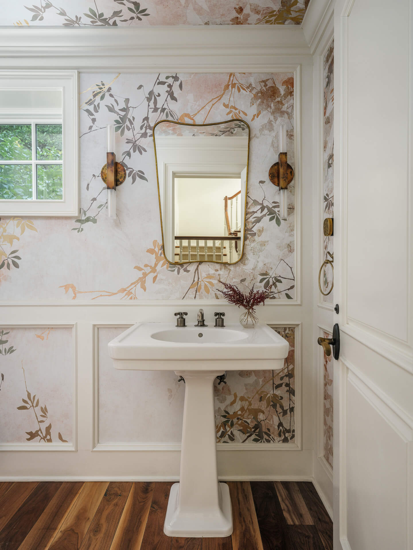 Powder room with floral wallpaper, white pedestal sink, ornate brass mirror, and dark wood floors.
