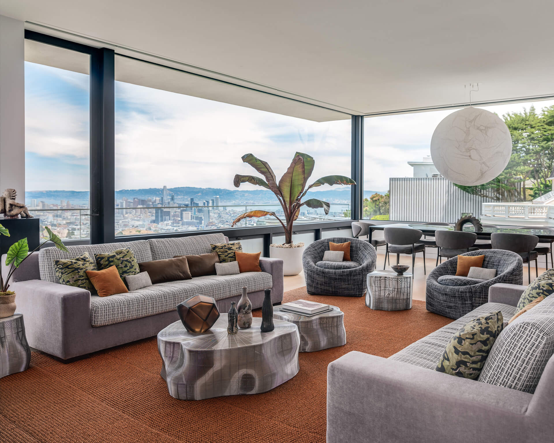 Spacious living room with layered neutral seating, sculptural tables, orange rug, and panoramic San Francisco views.