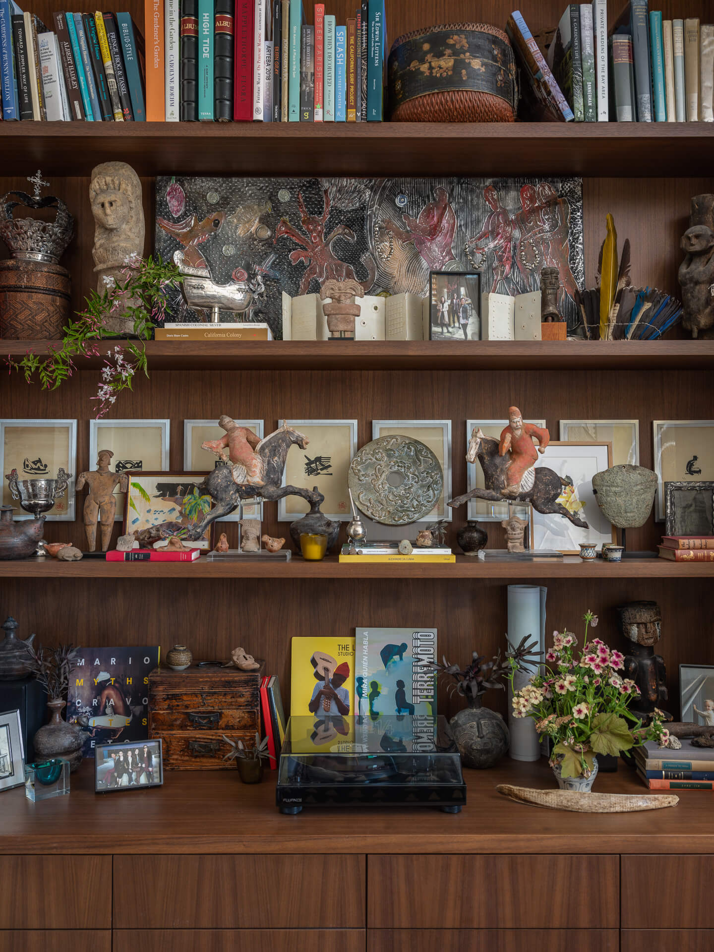 Wood display shelves densely arranged with books, ceramics, artwork, flowers, and collected objects.