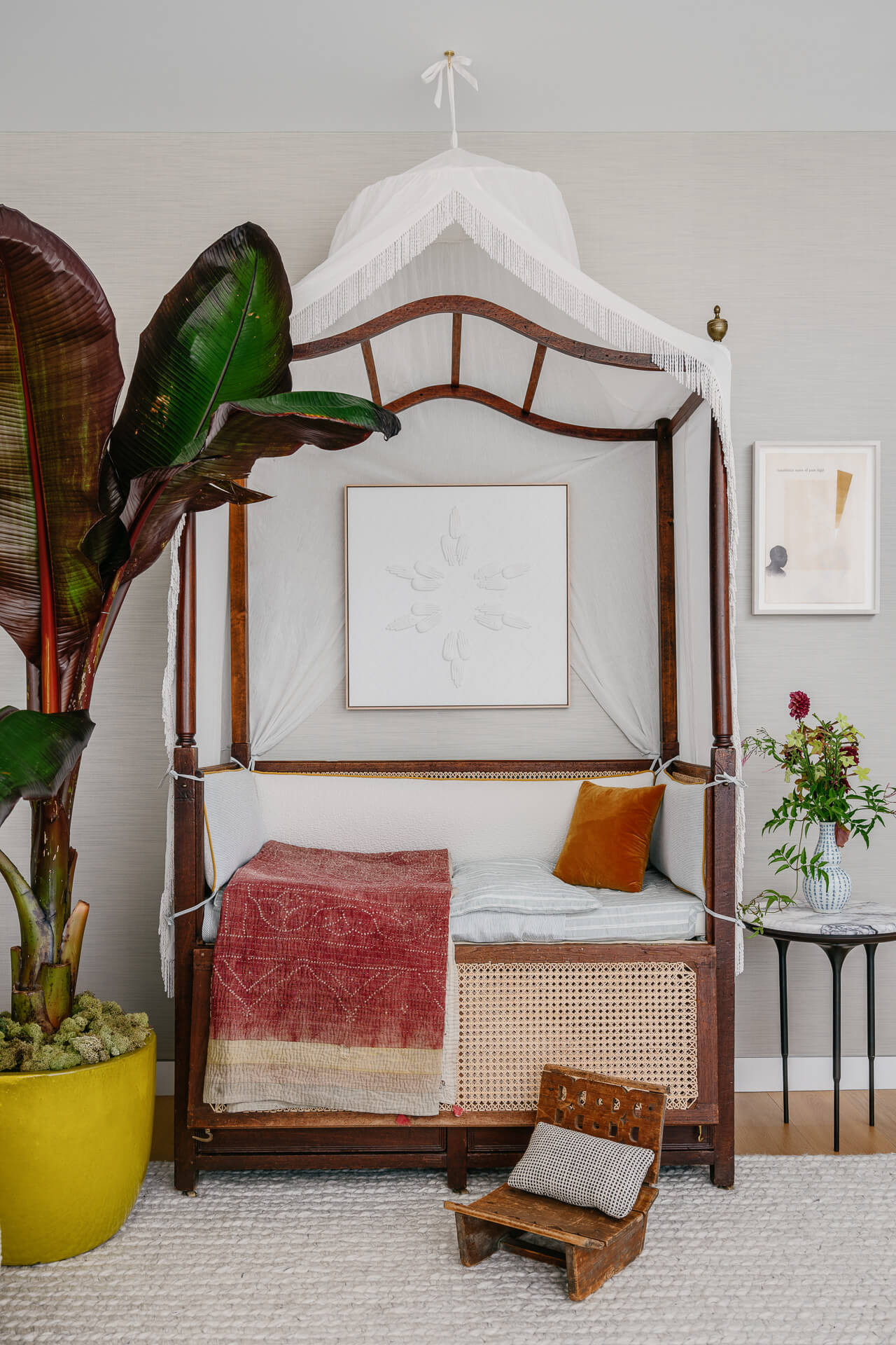 Decorative daybed with canopy frame, large tropical plant, red throw, and a miniature wood chair.