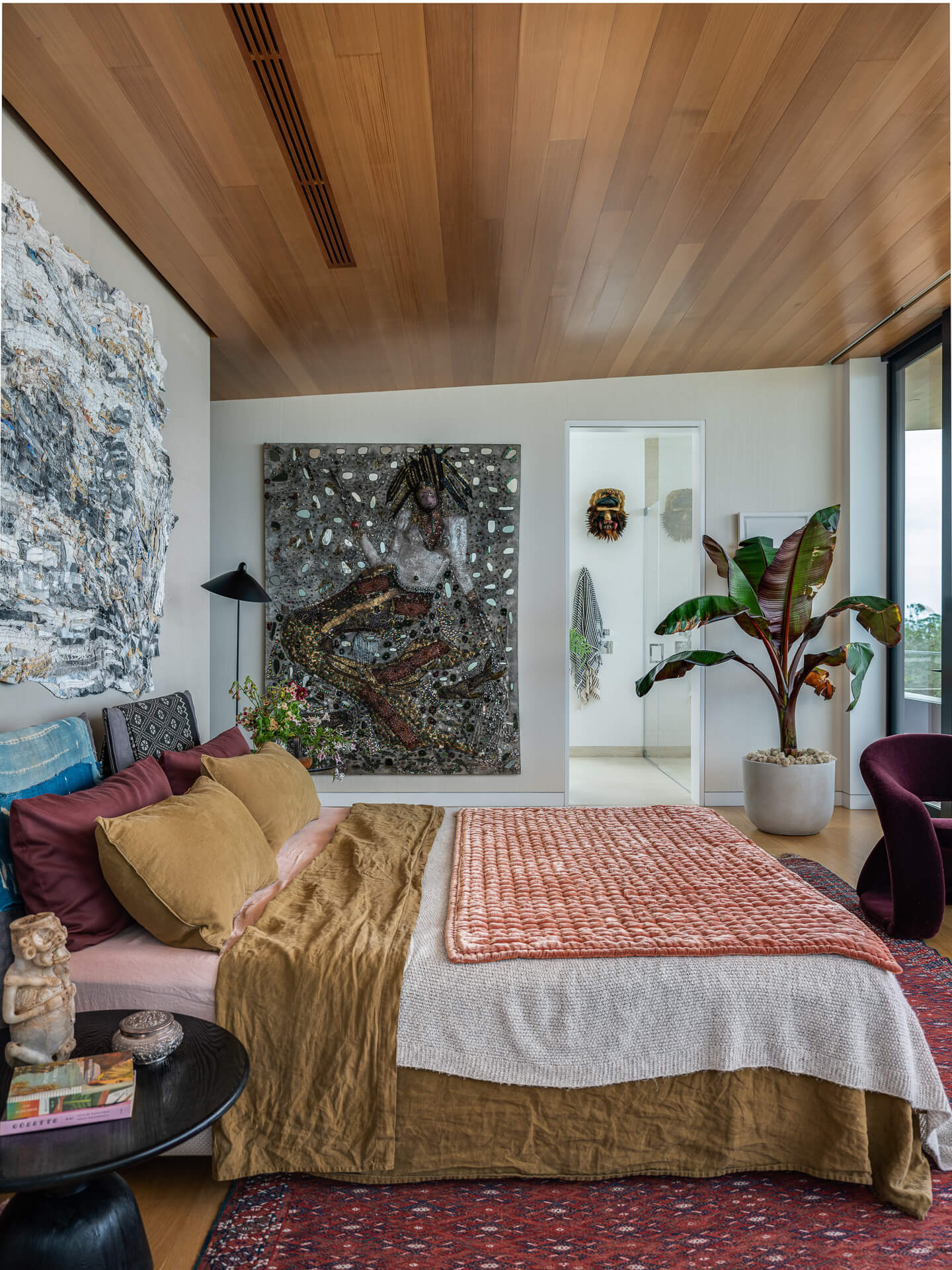 Bedroom with wood-paneled ceiling, layered bedding, colorful artwork, and floor-to-ceiling windows.