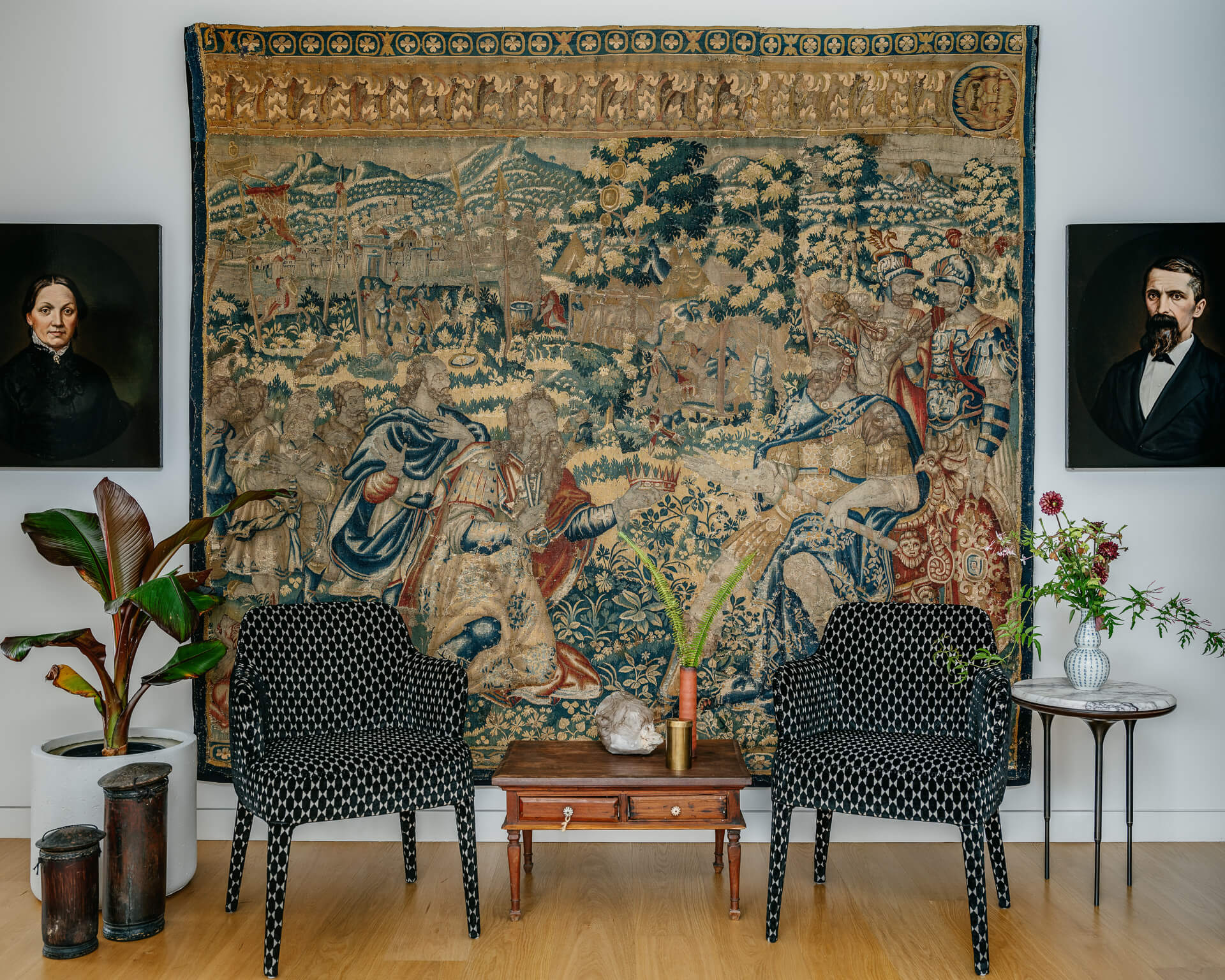 Two patterned armchairs beneath a large antique tapestry, flanked by portrait paintings and potted plants.
