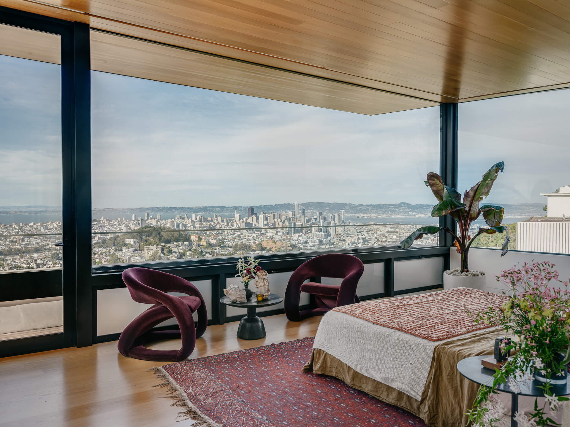 Bedroom with floor-to-ceiling corner windows, patterned rug, purple lounge chairs, and sweeping city views.