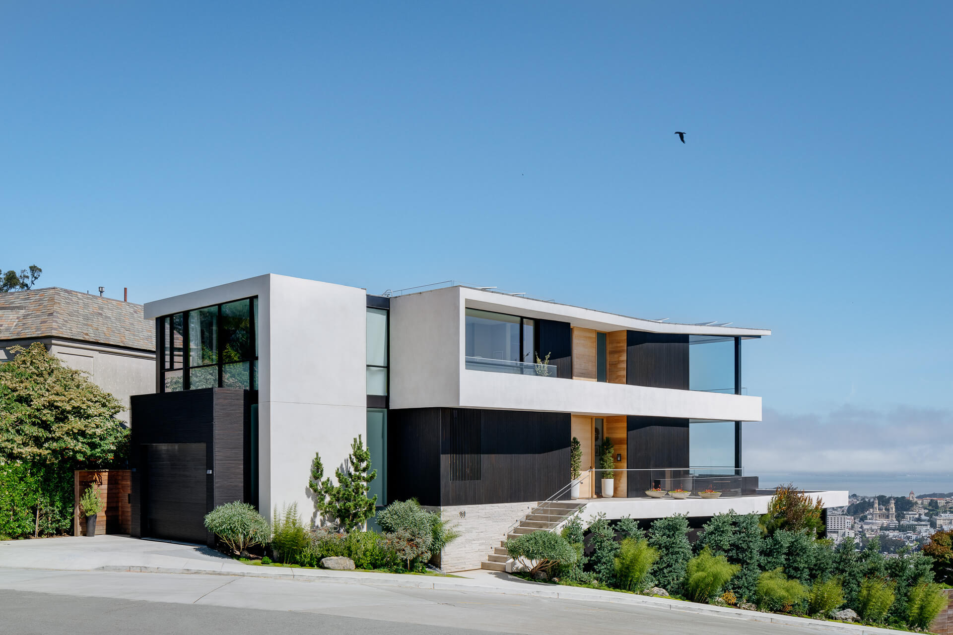 Modern hillside home with white and black facade, broad windows, and landscaped front garden.