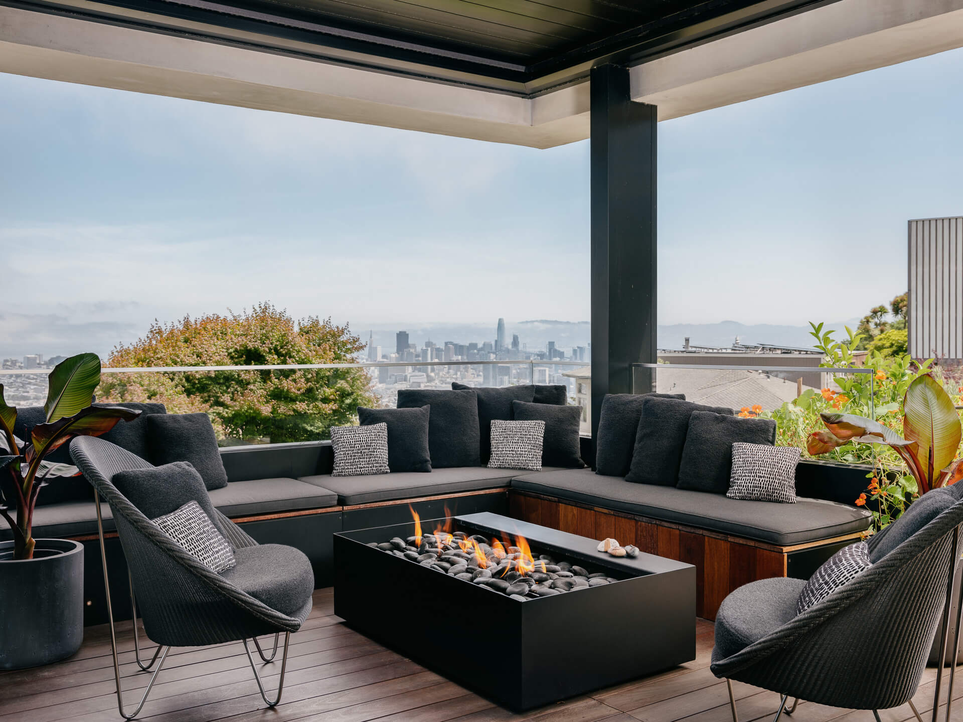 Covered outdoor lounge with dark sectional seating surrounding a rectangular fire pit and panoramic city views.