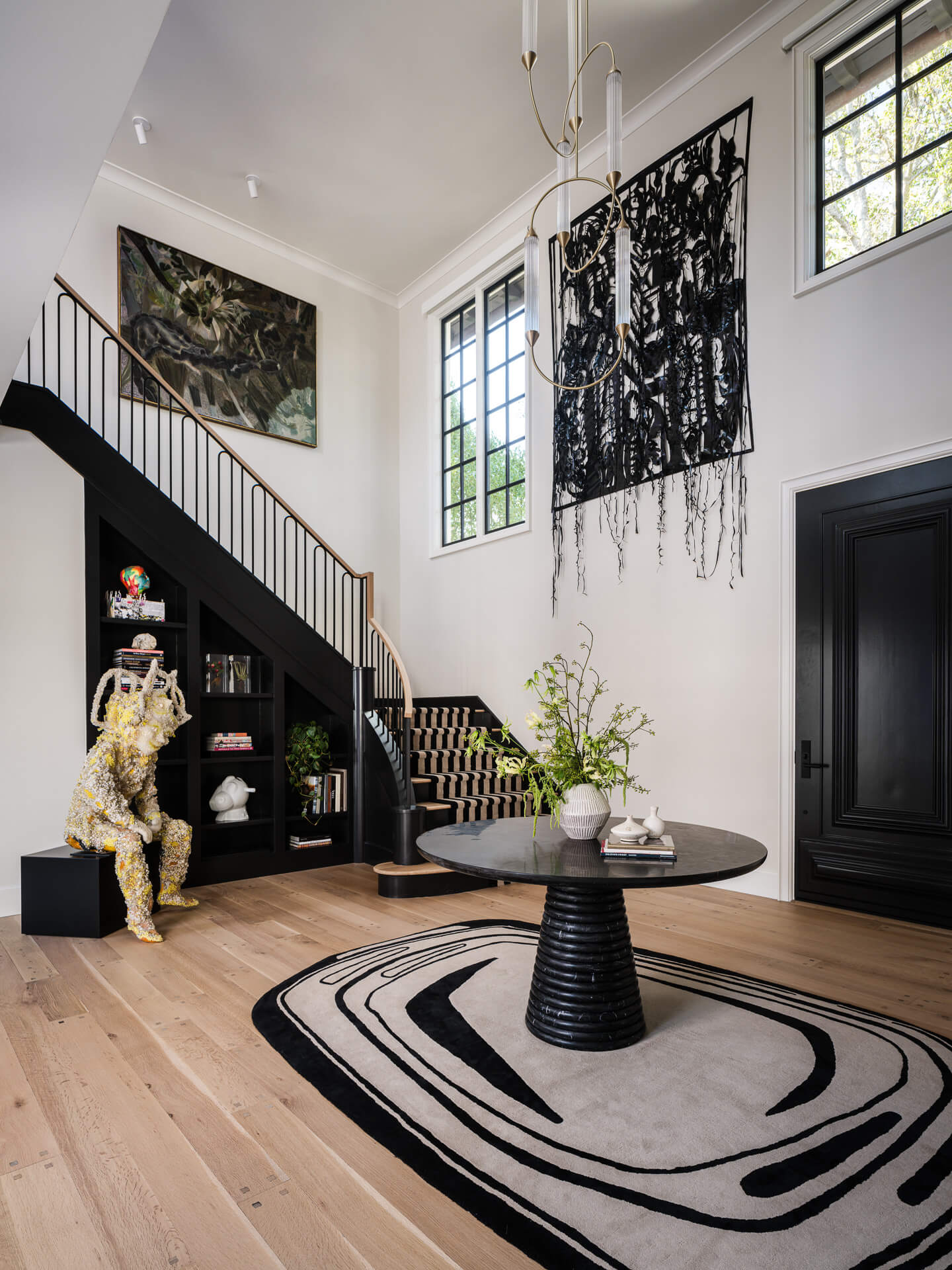 Double-height entry with black staircase, oversized artwork, sculptural furnishings, and light wood floors.