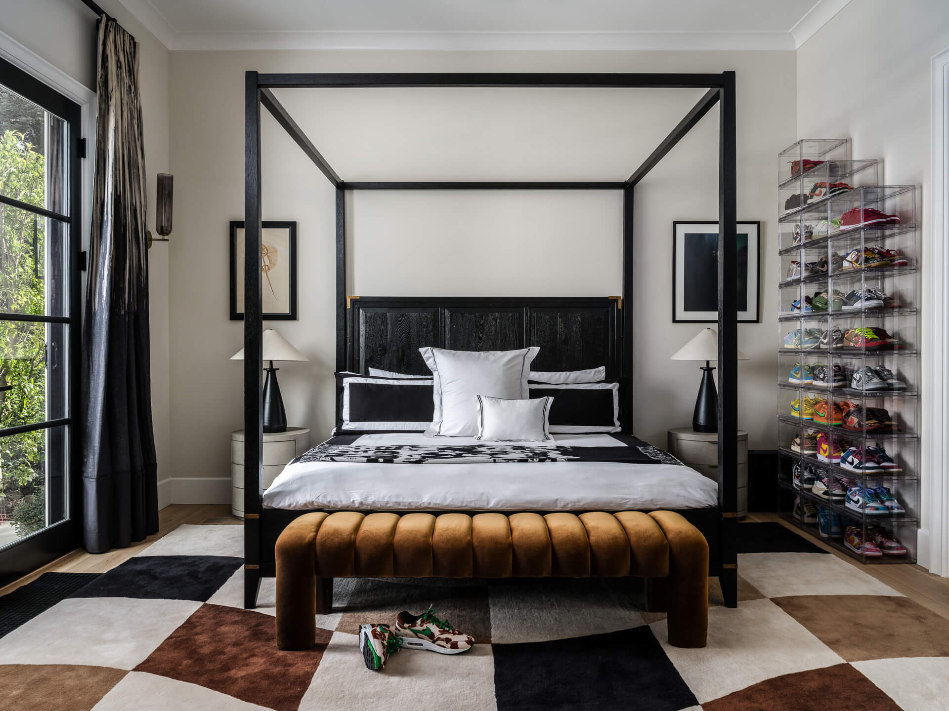 Bedroom with black canopy bed, caramel leather bench, geometric rug, and colorful shoe display.