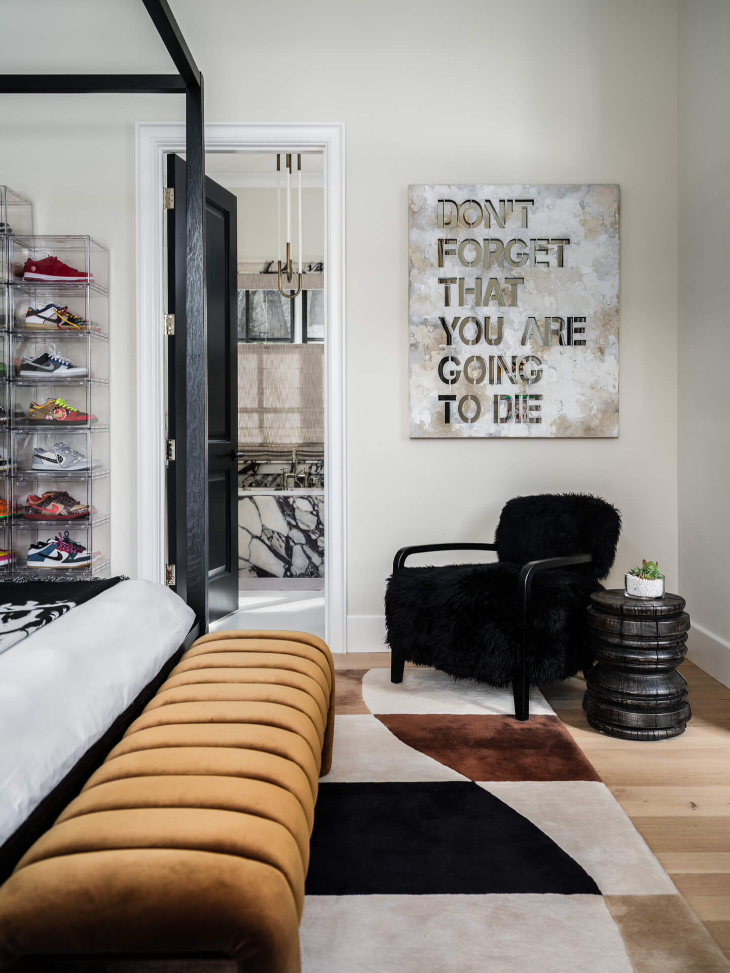 Bedroom corner with caramel bench, black lounge chair, stacked side table, and graphic wall art.