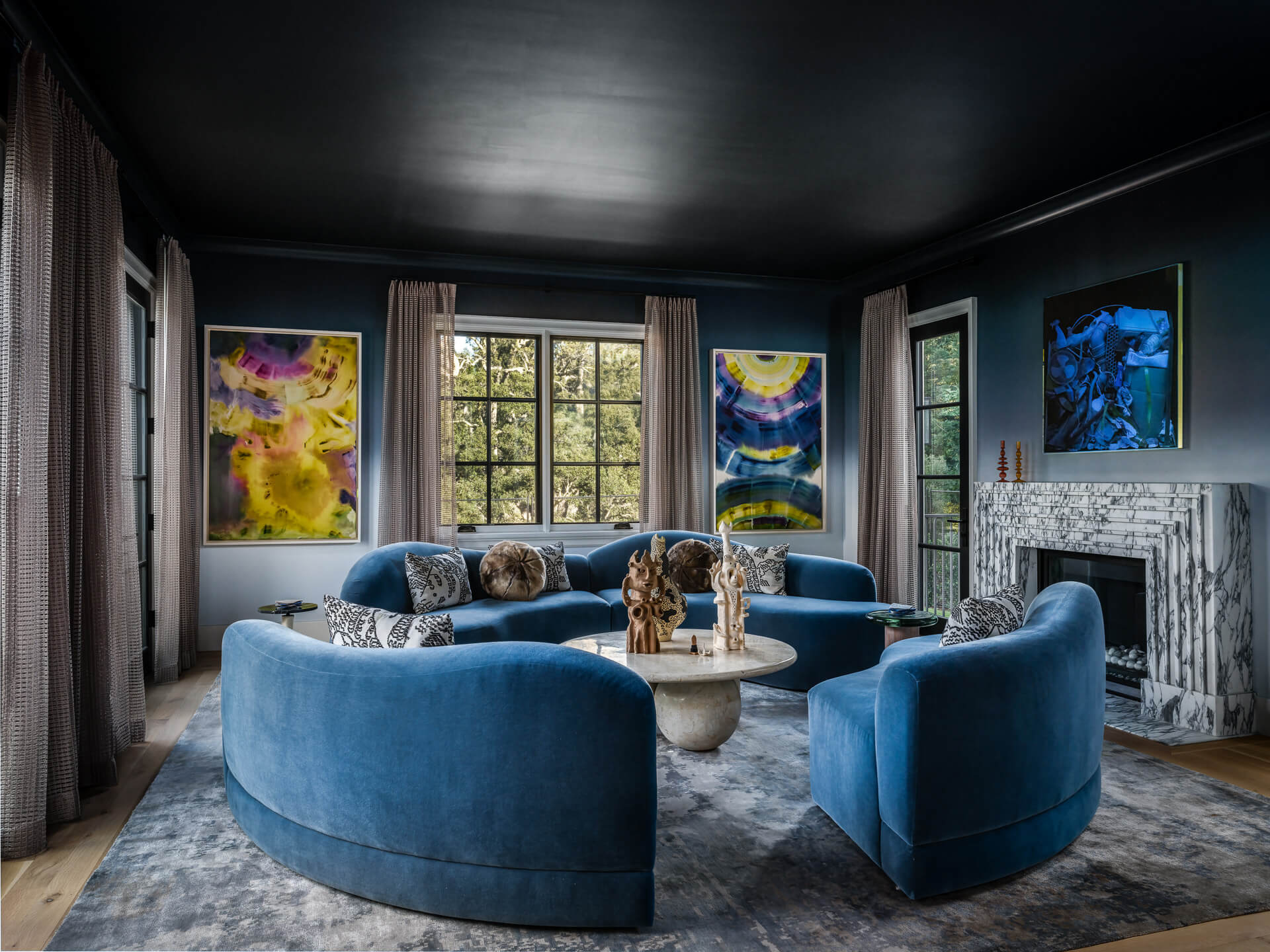 Moody living room with curved blue velvet seating, colorful artwork, dark ceiling, and marble fireplace.