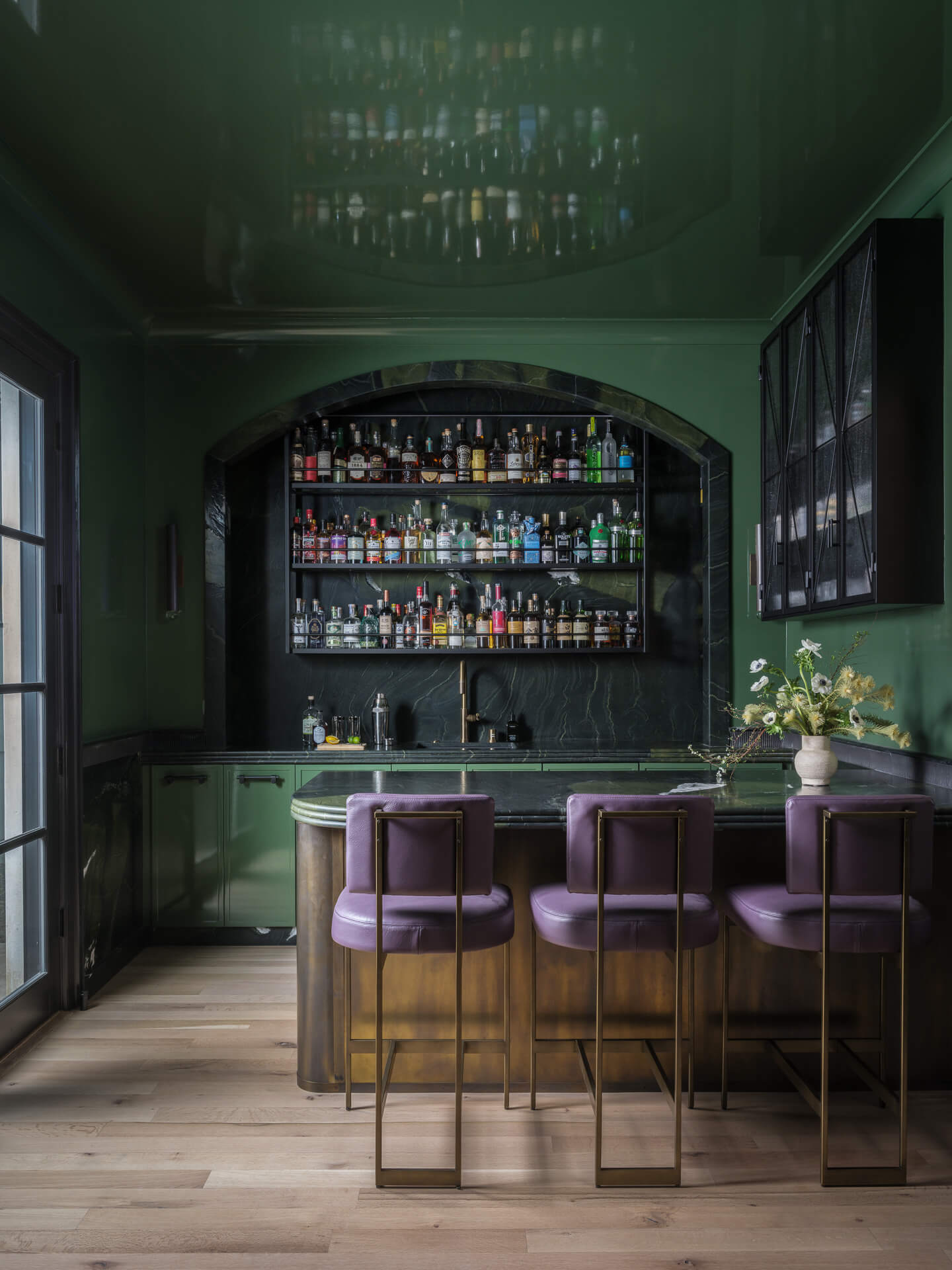 Deep green home bar with marble counter, purple stools, glass shelving, and chandelier overhead