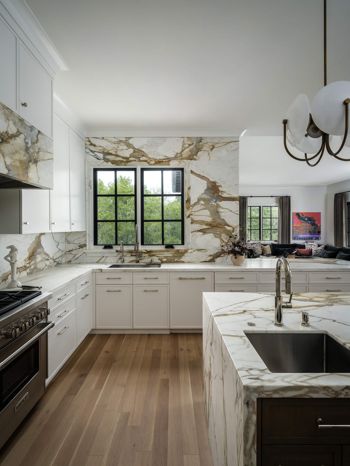 White kitchen with veined marble countertops and backsplash, black-framed window, and light wood floors.