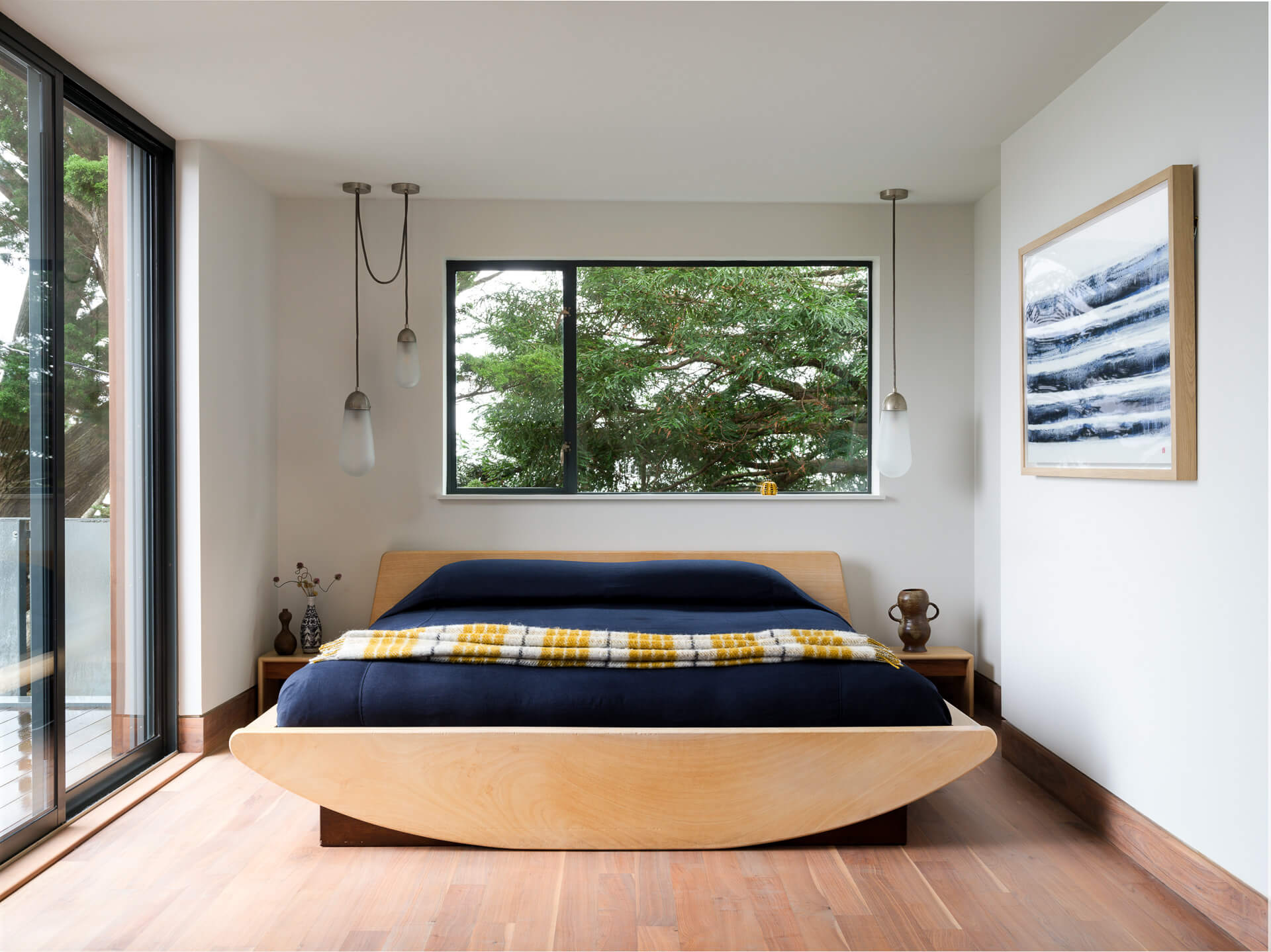 Minimal bedroom with low curved wood bed, dark bedding, and corner windows overlooking trees.