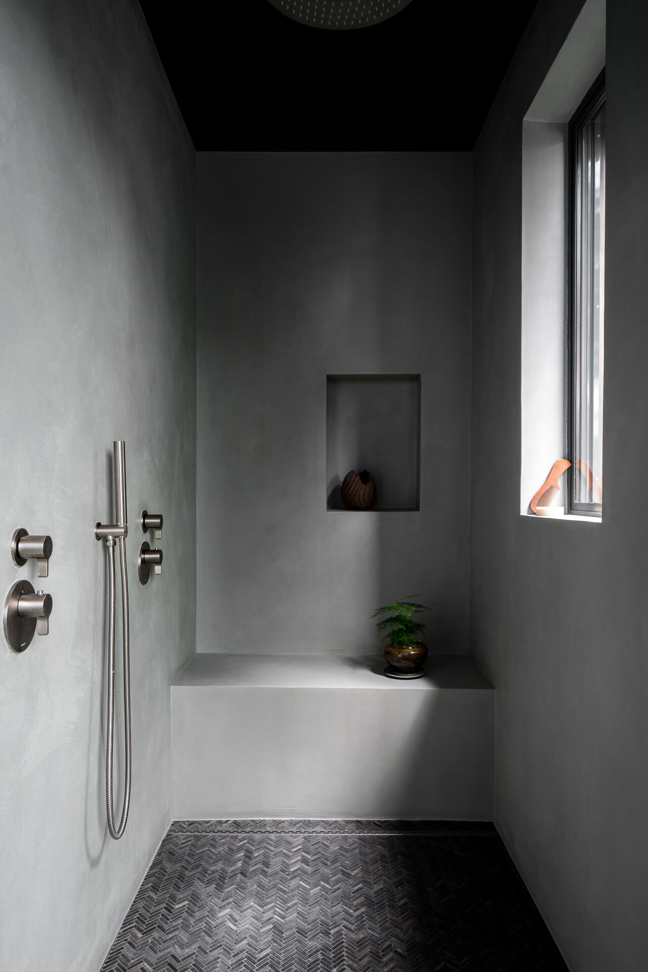 Minimal gray plaster shower with built-in bench, pebble floor, handheld fixture, and small window.