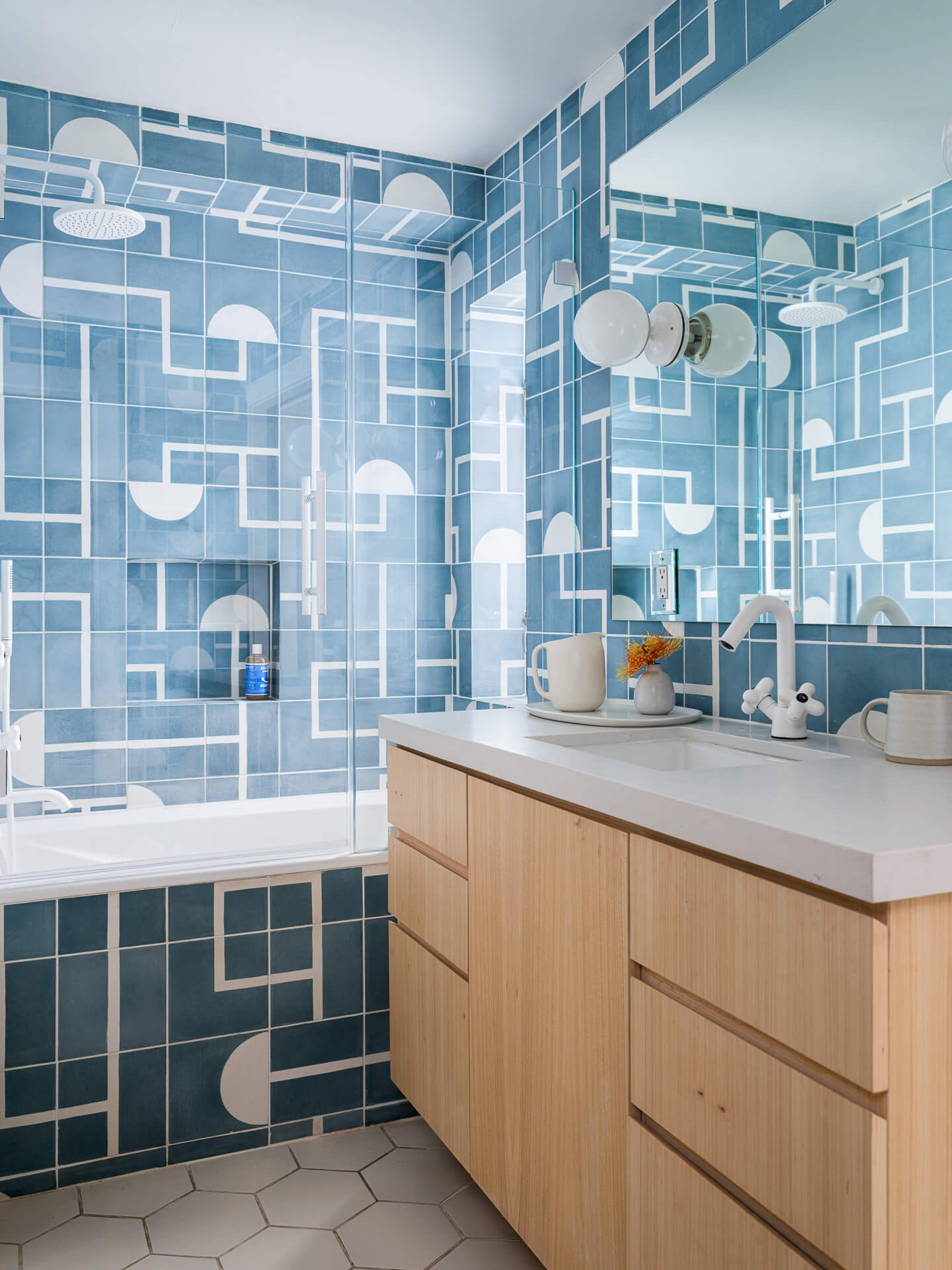 Blue patterned bathroom with light wood vanity, round mirrors, globe lights, and tiled tub surround.