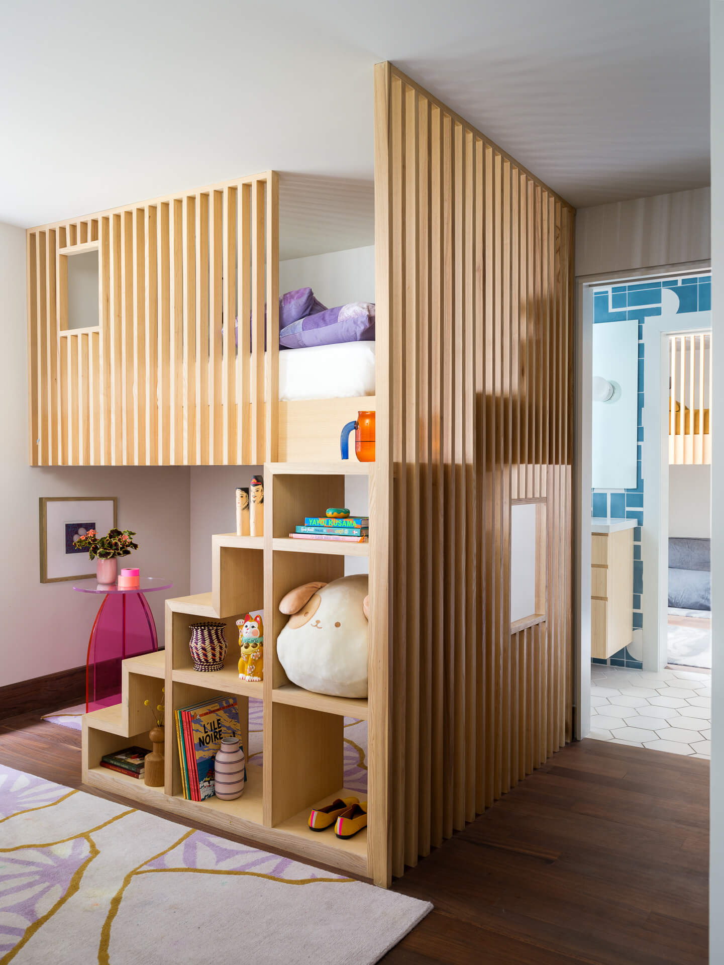 Children’s room with wood-slatted loft bed, open storage, built-in shelving, and small pink desk.