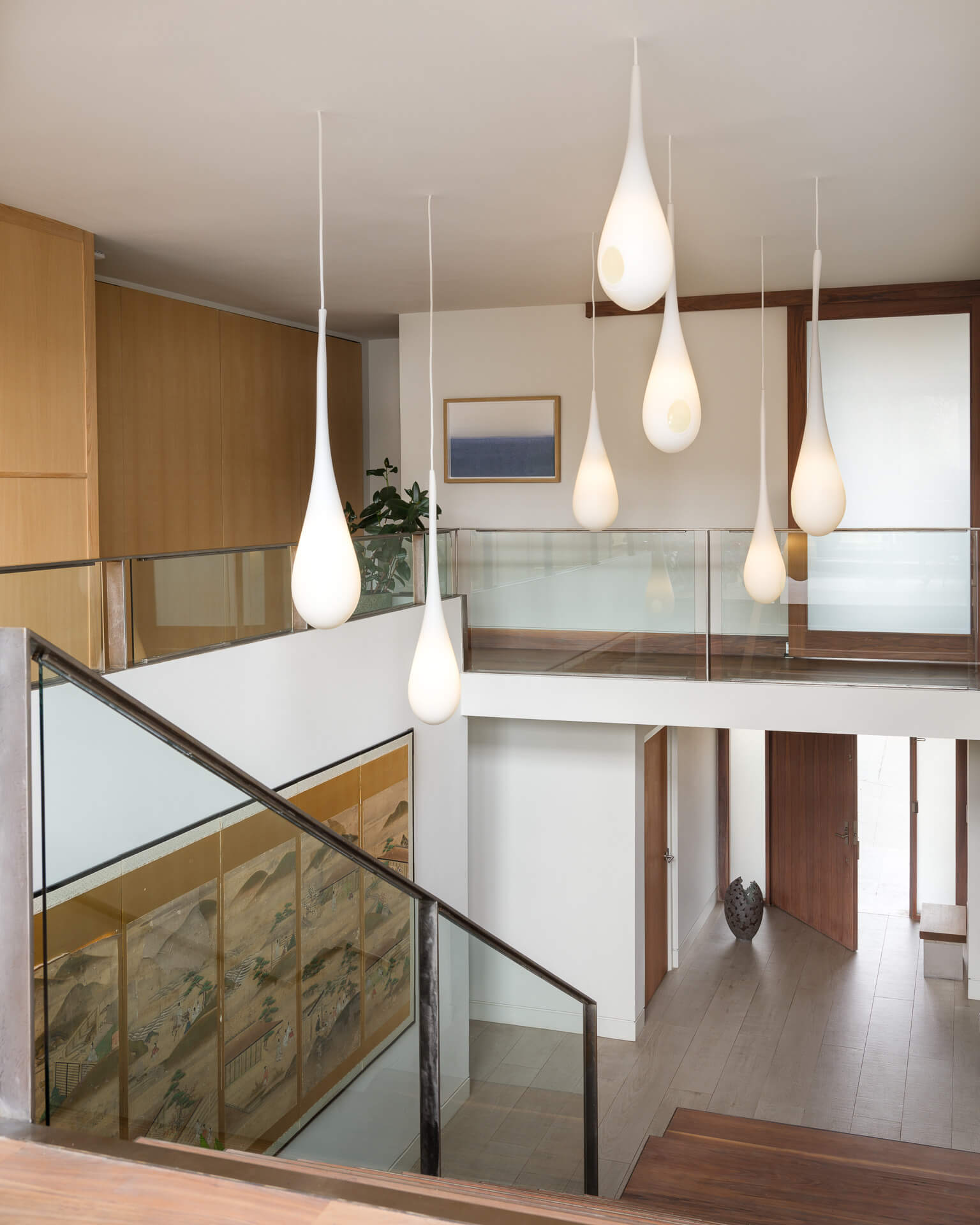 Open stair landing with glass railing, light wood cabinetry, and a cluster of white pendant lights.