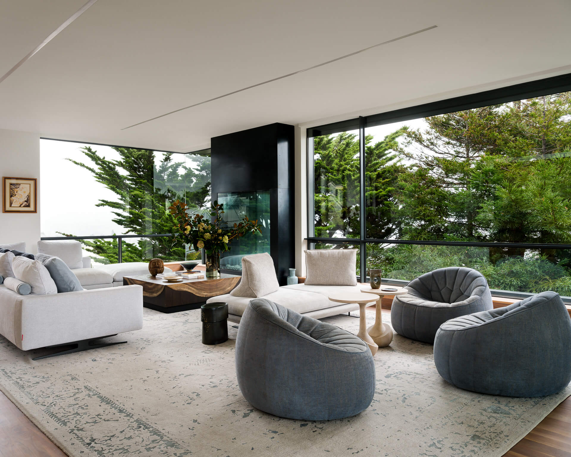 Bright living room with white sectional, rounded gray chairs, and floor-to-ceiling windows overlooking trees.